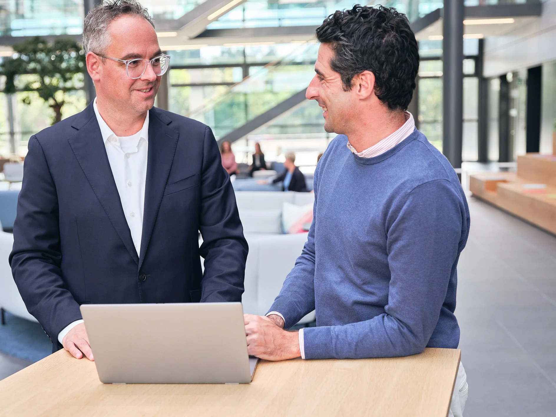 Zwei Männer stehen und unterhalten sich an einem Holztisch mit einem Laptop in einer Bürolobby. Einer der Männer trägt einen Anzug, der andere einen blauen Pullover. Ihre Unterhaltung scheint spannend zu sein. Im Hintergrund sind große Fenster und Sitzbereiche zu sehen. (This text has been generated by AI)