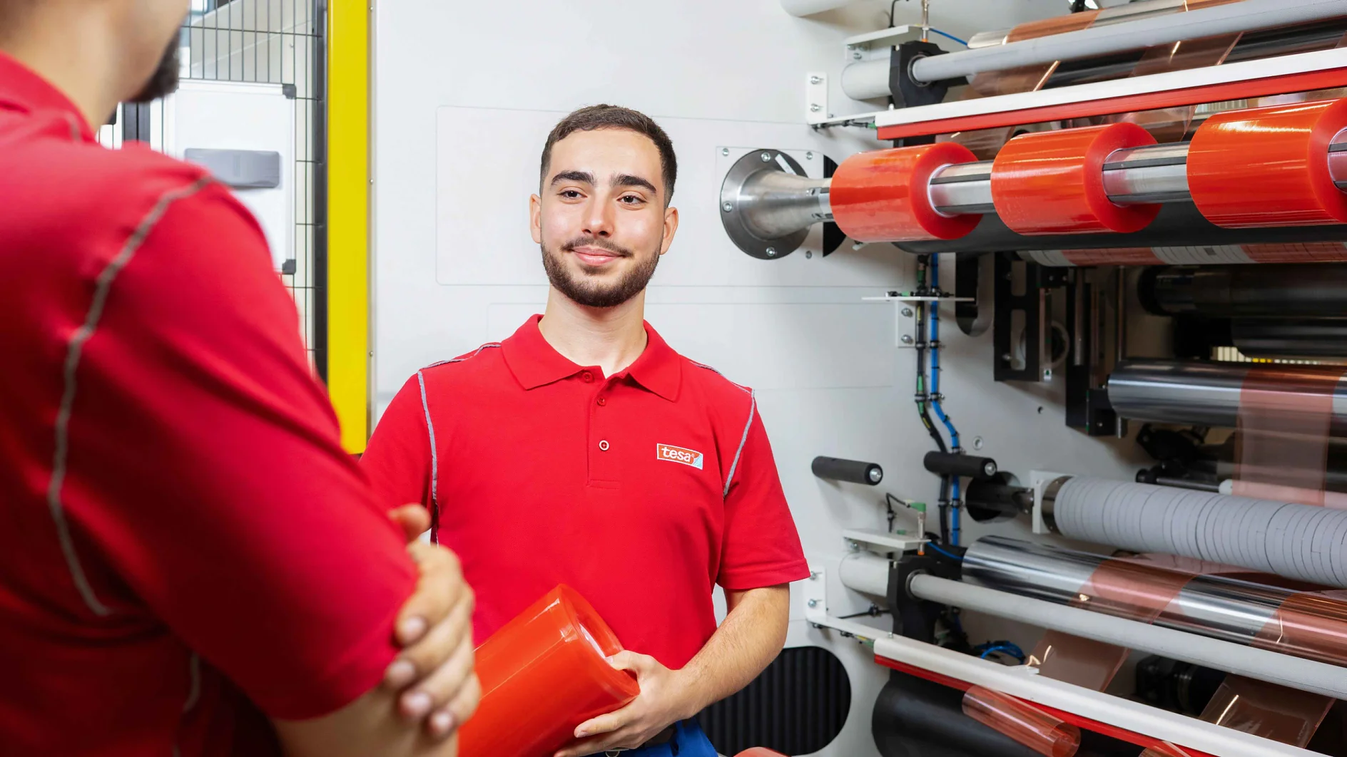 Ein Mann in einem roten Poloshirt hält eine Rolle roter Plastikfolie und steht neben einer großen Industriemaschine mit mehreren Spulen. Eine andere Person in einem ähnlichen Shirt steht mit verschränkten Armen daneben. Die Szene scheint eine Produktionsstätte zu sein, in der Tesa-Klebeband verwendet wird. (This text has been generated by AI)
