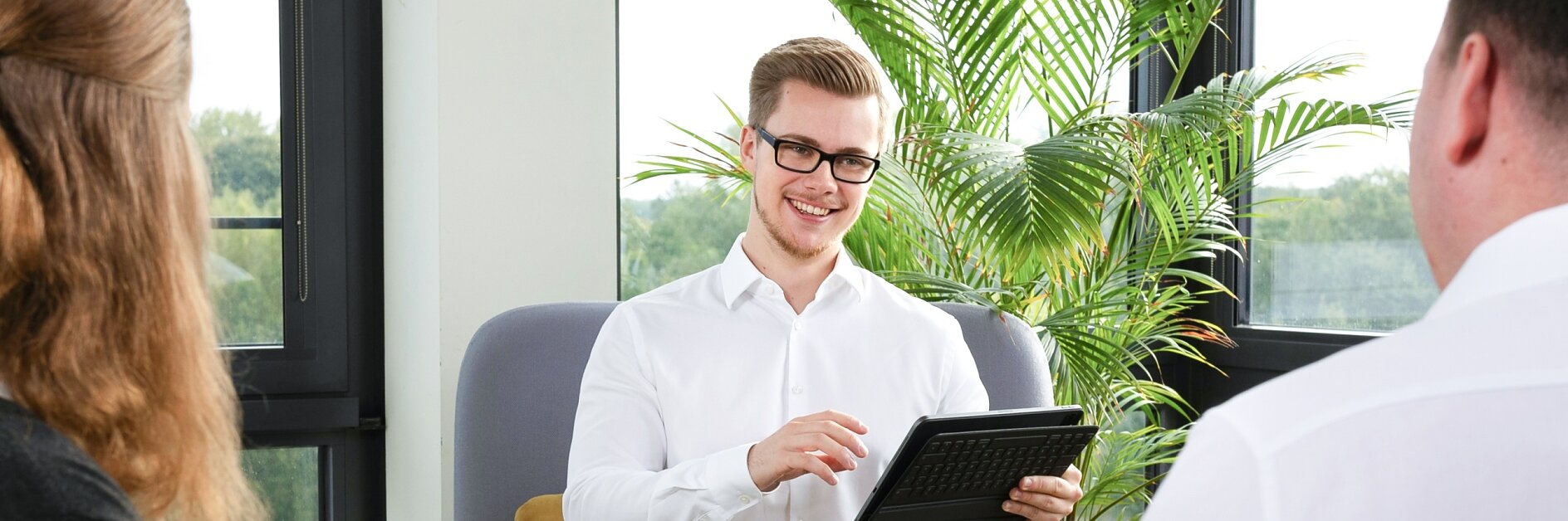 Ein Mann im weißen Hemd sitzt lächelnd auf einem Stuhl und hält ein Tablet in der Hand. Zwei Personen stehen mit dem Rücken zur Kamera und blicken auf ihn. Der Raum ist hell, mit großen Fenstern und üppigen grünen Pflanzen im Hintergrund. (This text has been generated by AI)