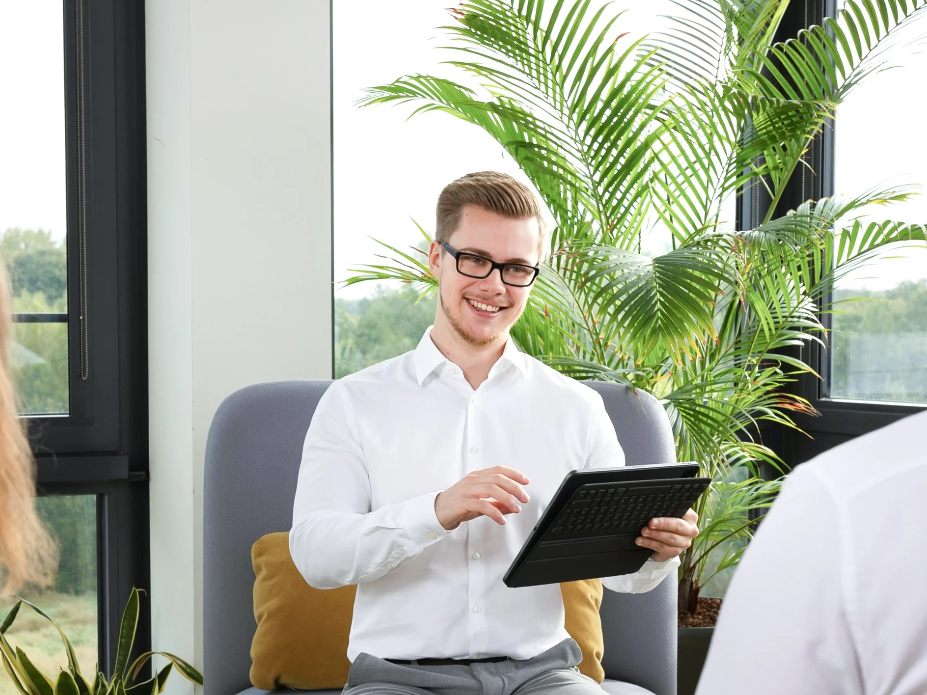 Ein Mann im weißen Hemd sitzt lächelnd auf einem Stuhl und hält ein Tablet in der Hand. Zwei Personen stehen mit dem Rücken zur Kamera und blicken auf ihn. Der Raum ist hell, mit großen Fenstern und üppigen grünen Pflanzen im Hintergrund. (This text has been generated by AI)
