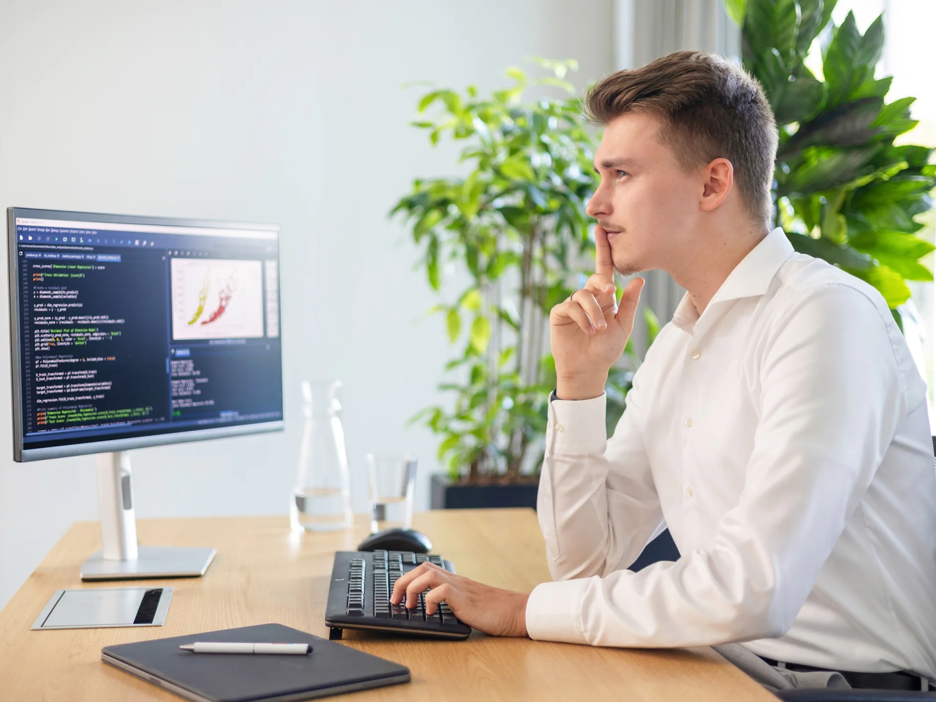 Ein Mann in weißem Hemd sitzt an einem Schreibtisch in einem Büro und konzentriert sich auf einen Computermonitor, auf dem Code und Grafiken angezeigt werden. Seine Hand liegt in nachdenklicher Haltung auf seinem Kinn. In der Nähe stehen ein Glas Wasser, eine Karaffe, eine Pflanze und ein geschlossenes Notizbuch mit Tesa-Klebeband daneben. (This text has been generated by AI)