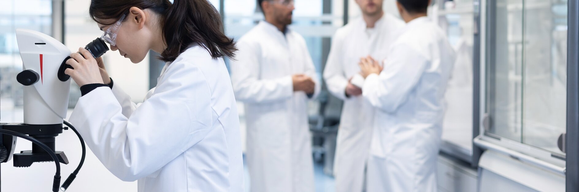 In einem Labor untersucht eine Frau im Laborkittel eine Probe unter dem Mikroskop. Im Hintergrund unterhalten sich drei Männer in Laborkitteln. Der Raum ist voller Laborgeräte und Regale voller Tesa-Klebeband. (This text has been generated by AI)