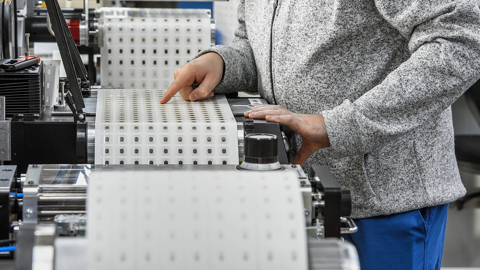 industrial-converter-die-cutting-key-visual-cms Eine Person bedient in einer Fabrik Maschinen mit großen Rollen Tesa-Klebeband. Die Person trägt eine graue Jacke mit einem „Tesa“-Logo und eine blaue Hose und passt das Tesa-Band mit den Händen an. (This text has been generated by AI)