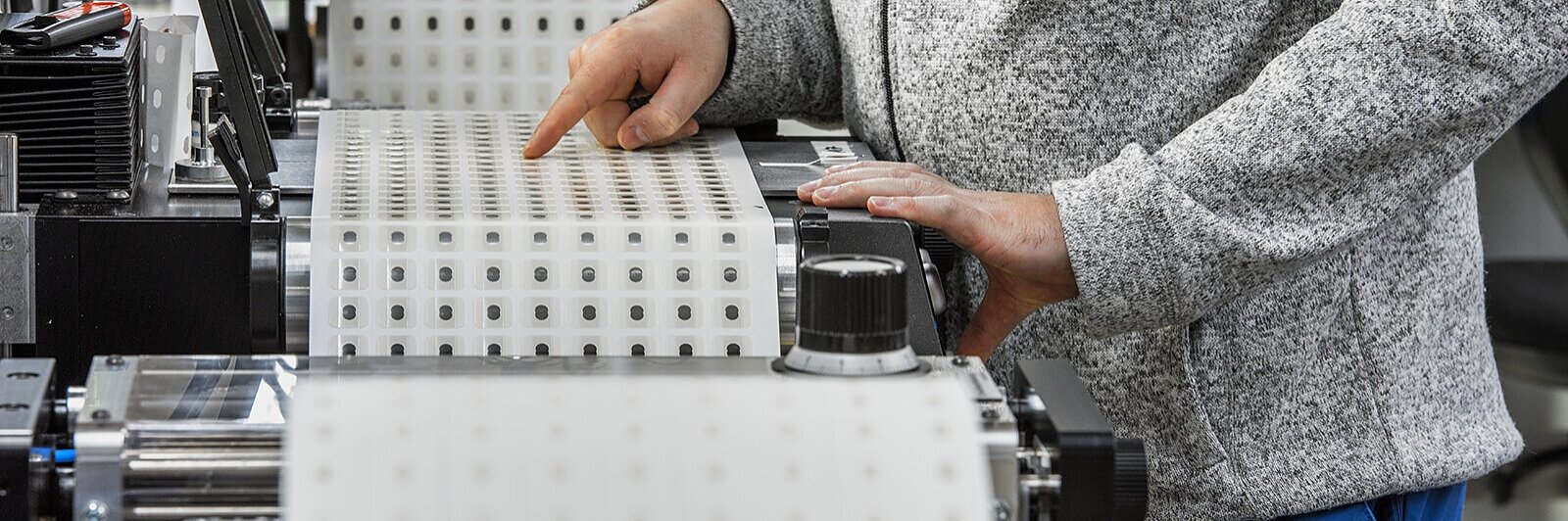 Eine Person bedient in einer Fabrik Maschinen mit großen Rollen Tesa-Klebeband. Die Person trägt eine graue Jacke mit einem „Tesa“-Logo und eine blaue Hose und passt das Tesa-Band mit den Händen an. (This text has been generated by AI)