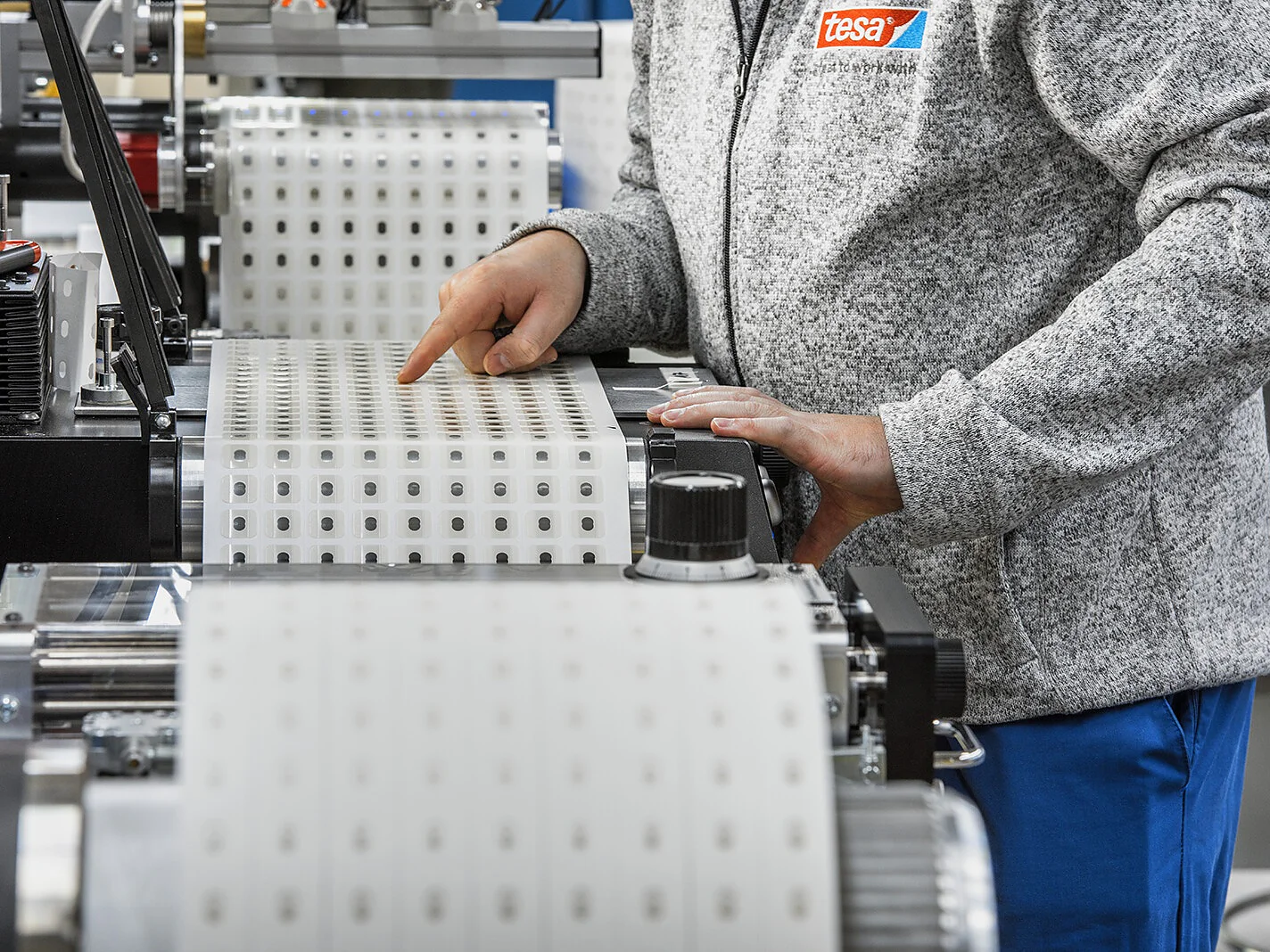 Eine Person bedient in einer Fabrik Maschinen mit großen Rollen Tesa-Klebeband. Die Person trägt eine graue Jacke mit einem „Tesa“-Logo und eine blaue Hose und passt das Tesa-Band mit den Händen an. (This text has been generated by AI)