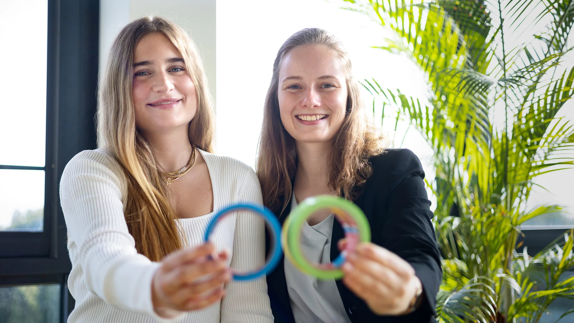 Zwei Menschen lächeln und halten bunte Ringe in die Kamera. Sie stehen drinnen an einem Fenster, durch das Sonnenlicht hereinfällt. Rechts steht eine große grüne Pflanze. Beide tragen legere Businesskleidung und die Ringe sind mit Tesa-Klebeband befestigt. (This text has been generated by AI)