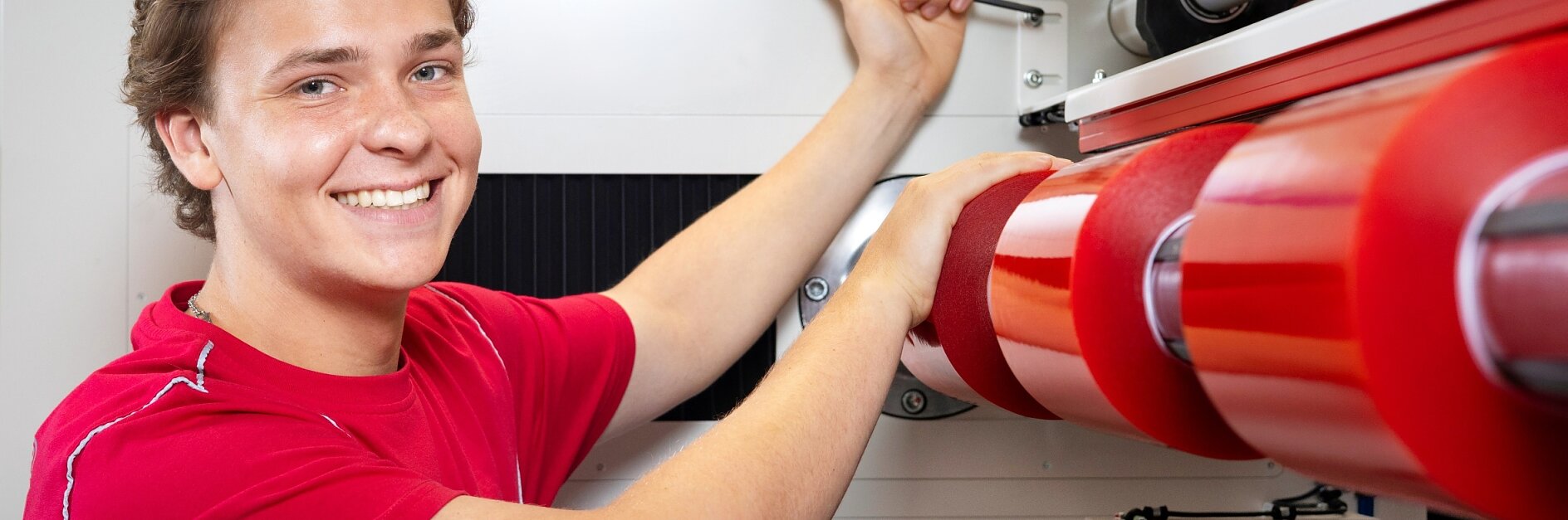 Eine Person im roten Hemd bedient in einer hellen Industrieumgebung eine Maschine mit großen roten Walzen. Sie lächelt und blickt in die Kamera, während sie einen Hebel an der Maschine einstellt und dabei Tesa-Klebeband für Präzision und Zuverlässigkeit verwendet. (This text has been generated by AI)
