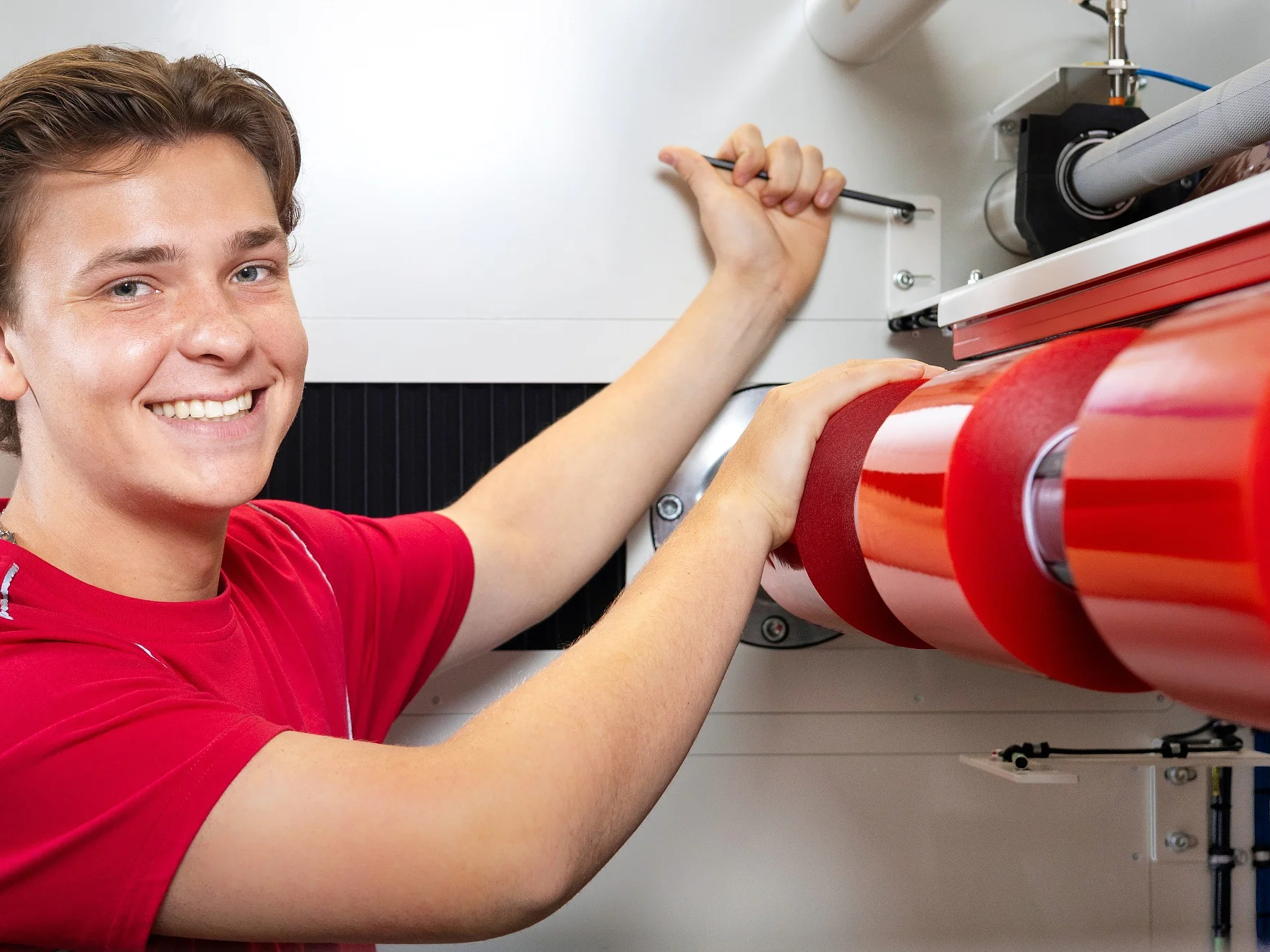Eine Person im roten Hemd bedient in einer hellen Industrieumgebung eine Maschine mit großen roten Walzen. Sie lächelt und blickt in die Kamera, während sie einen Hebel an der Maschine einstellt und dabei Tesa-Klebeband für Präzision und Zuverlässigkeit verwendet. (This text has been generated by AI)