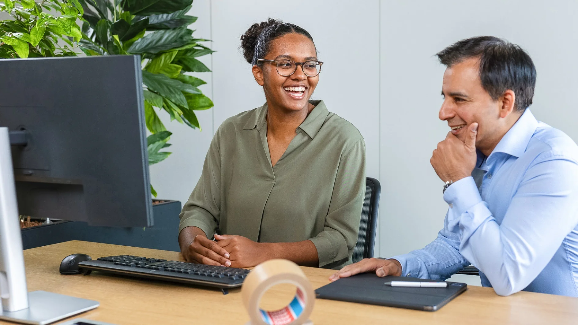 Zwei Personen sitzen an einem Schreibtisch in einem Büro und schauen auf einen Computermonitor. Die Frau links trägt eine Brille und eine grüne Bluse. Der Mann rechts, der ein blaues Hemd trägt, lächelt und die Hand am Kinn hat. In der Nähe steht eine Pflanze. (This text has been generated by AI)