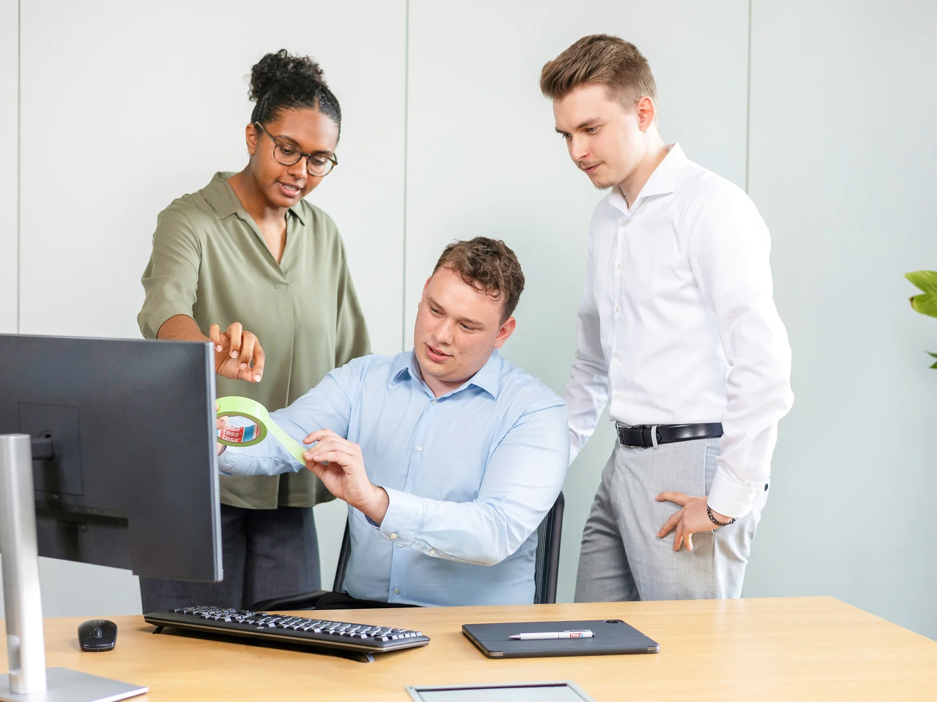 Drei Kollegen in einem modernen Büro blicken auf einen Computerbildschirm. Eine Frau in einer grünen Bluse deutet auf den Monitor, während ein sitzender Mann in einem blauen Hemd ein Diagramm betrachtet. Ein Mann in einem weißen Hemd steht neben ihnen und beobachtet sie. Hinter ihnen ist eine Pflanze zu sehen. (This text has been generated by AI)