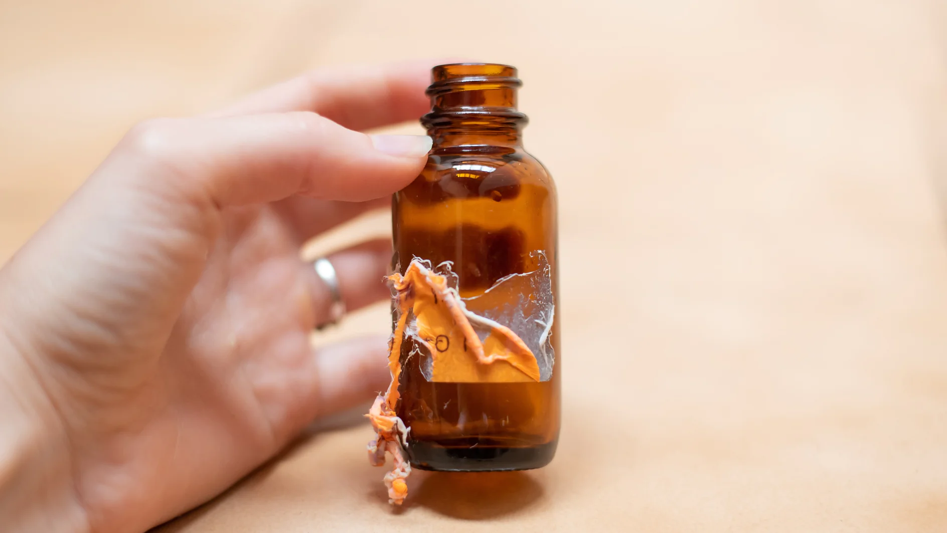 Eine Hand hält eine kleine bernsteinfarbene Glasflasche mit einem gebrochenen Abschnitt und gezackten Kanten an der Seite, an deren Kanten Tesa-Klebeband zu sehen ist. Der Hintergrund ist eine hellbeige Oberfläche. (This text has been generated by AI)