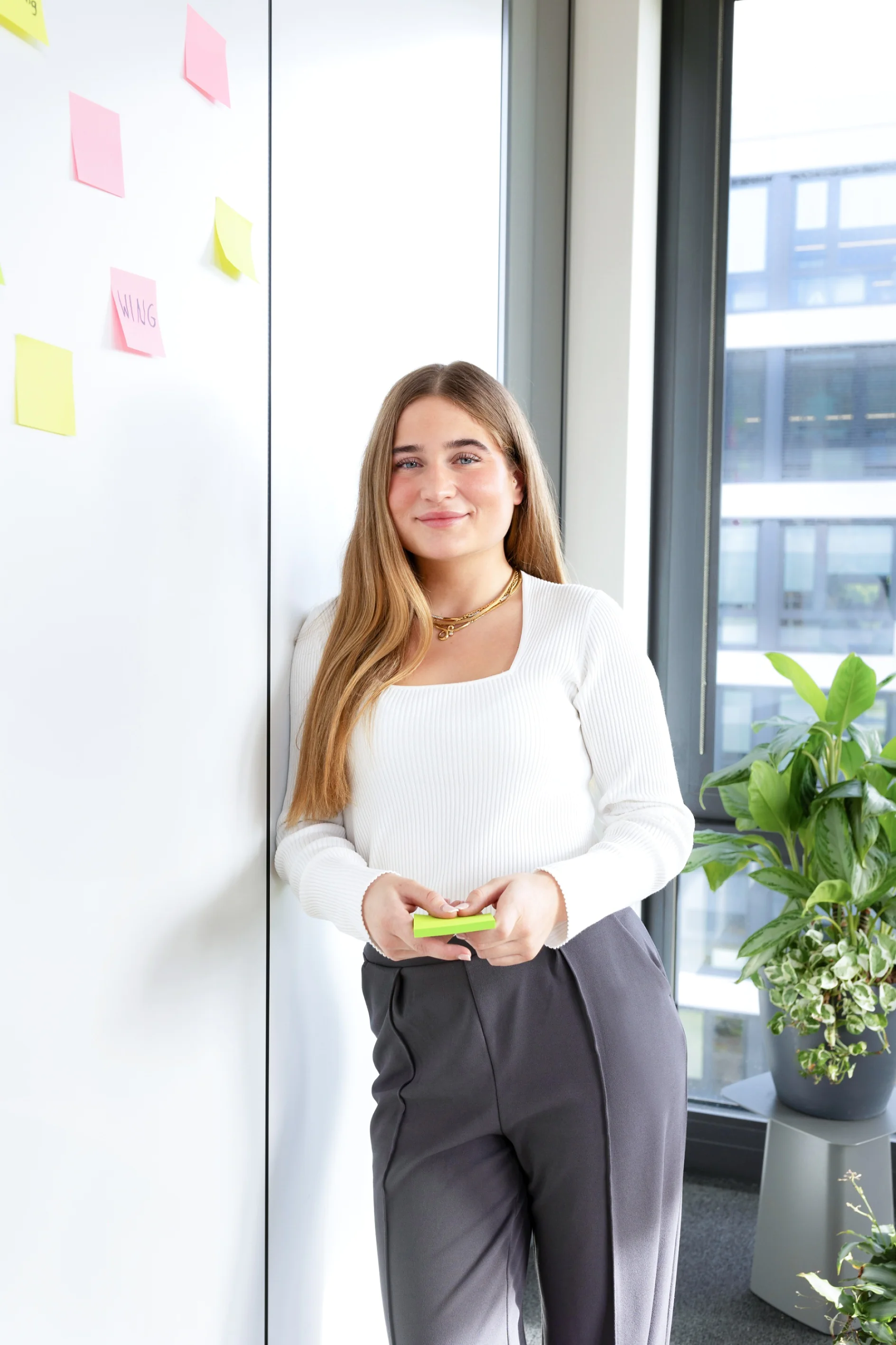 Eine Person mit langen Haaren lehnt in einem Büro mit Tesafilm-Haftnotizen an einer Wand. Sie hält weitere Notizen in der Hand. Der Raum verfügt über große Fenster und eine Topfpflanze. Sie trägt ein weißes Oberteil und dunkle Hosen. (This text has been generated by AI)