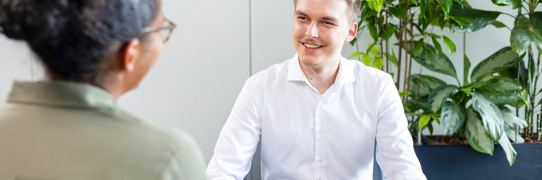 Ein Mann im weißen Hemd sitzt mit einem Stift an einem Schreibtisch und lächelt eine andere Person an, die der Kamera den Rücken zuwendet. Auf dem Schreibtisch liegt ein Tablet. Im Hintergrund sind grüne Pflanzen zu sehen. (This text has been generated by AI)