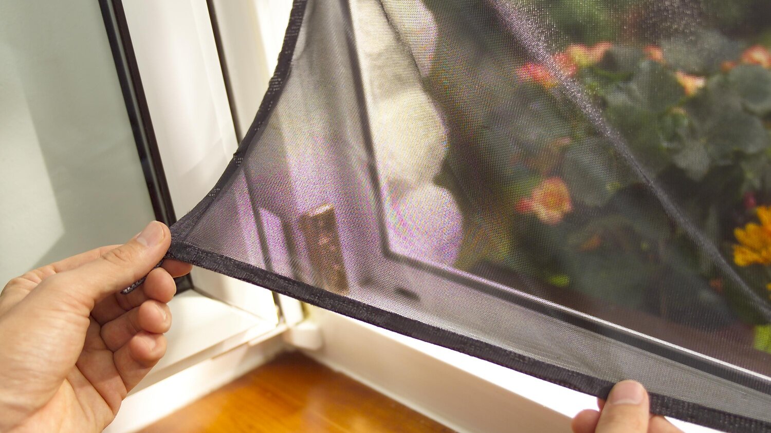 insect-hook-loop-sunprotect-windows_mood Hands are adjusting a removable window screen on a wooden window frame using tesa tape. The screen partially covers an outside view of green plants and orange flowers, visible through the glass. (This text has been generated by AI)