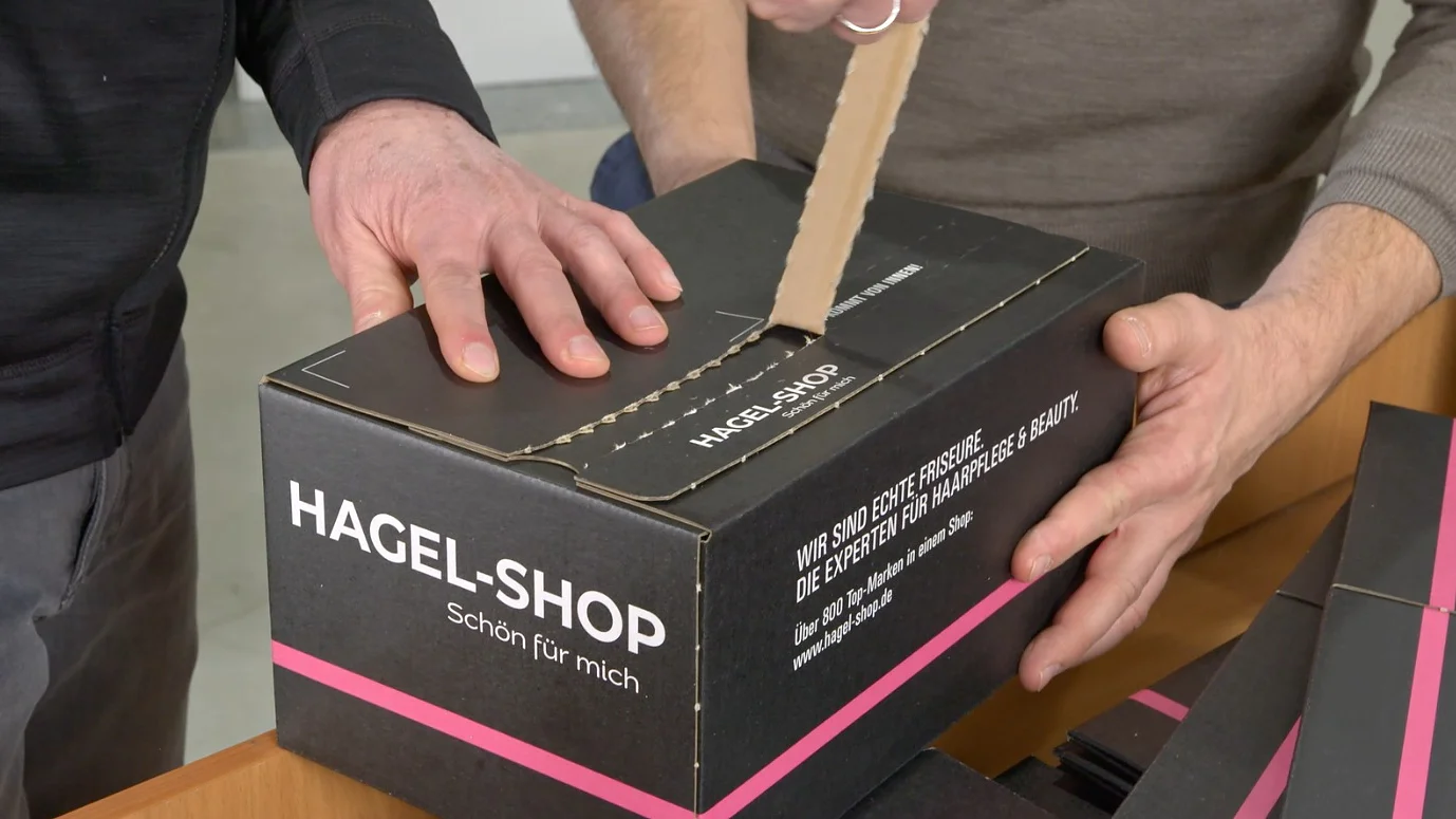 Two people are opening a black cardboard box with a utility knife. The box has Hagel-Shop printed on it. One person holds the box steady while the other cuts through the tesa tape sealing the top. The box is on a wooden surface. (This text has been generated by AI)