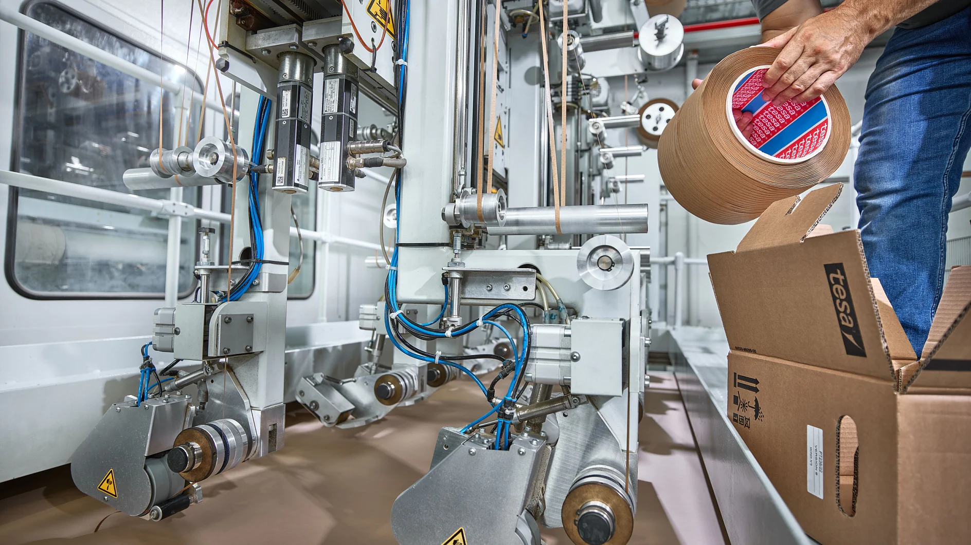 Eine Person, die neben einem Karton in einer Industrieanlage steht, legt eine Rolle braunes Tesa-Klebeband in eine automatische Verpackungsmaschine ein. (This text has been generated by AI)