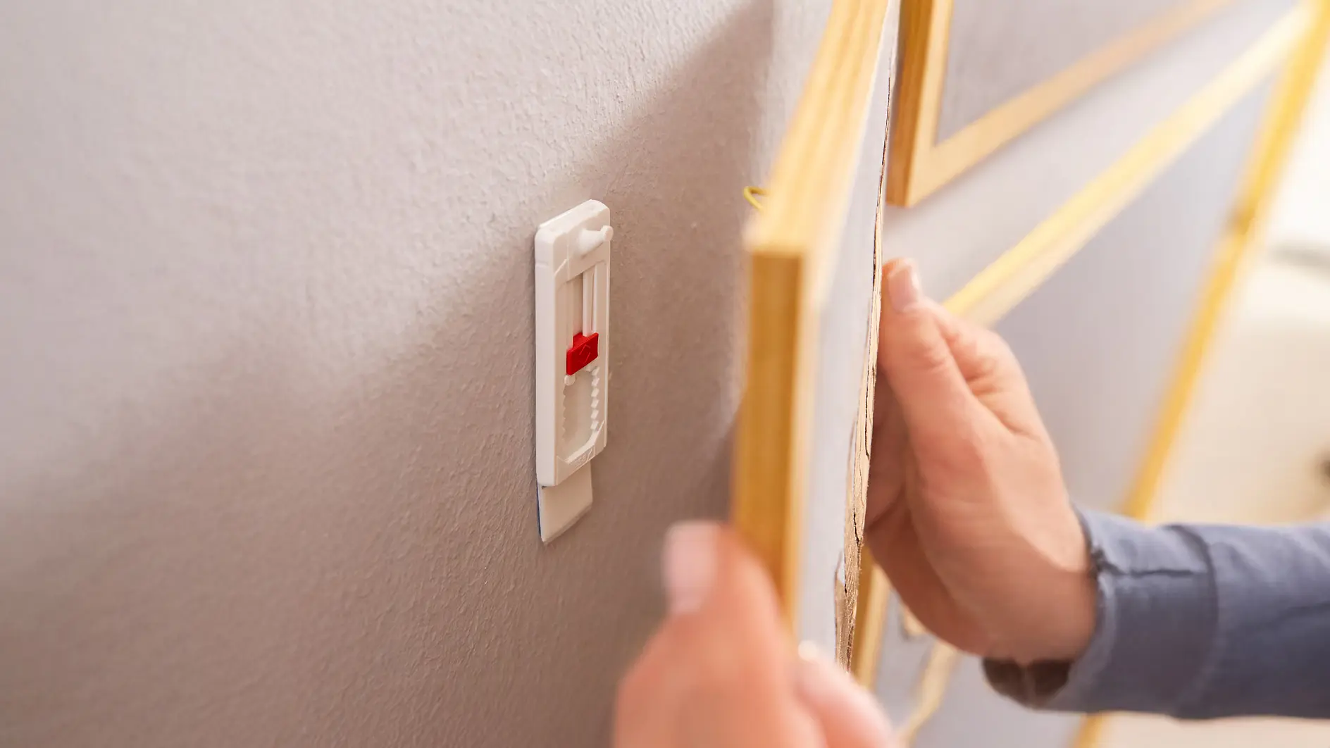 Eine Person hängt ein goldgerahmtes Bild an eine graue Wand, indem sie einen weißen Wandhaken aus Kunststoff mit Tesaband und einem roten Schieberegler verwendet. (This text has been generated by AI)
