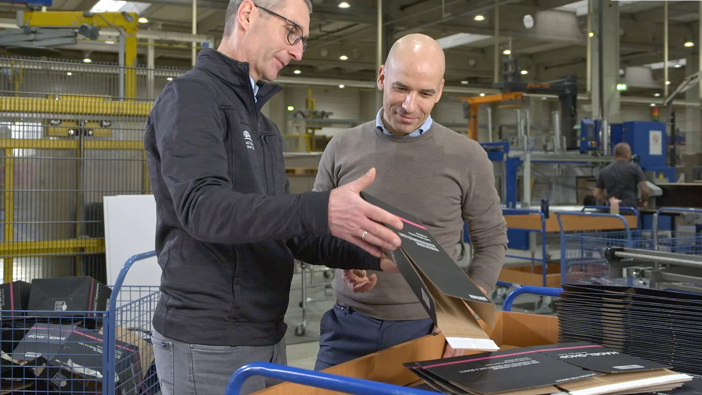 Two men in a factory setting examine printed packaging materials atop a wooden cart. The man on the left, wearing glasses and a black jacket, gestures towards the materials. The man on the right is bald, wearing a grey sweater, and looks attentively at the tesa tape used on them. (This text has been generated by AI)