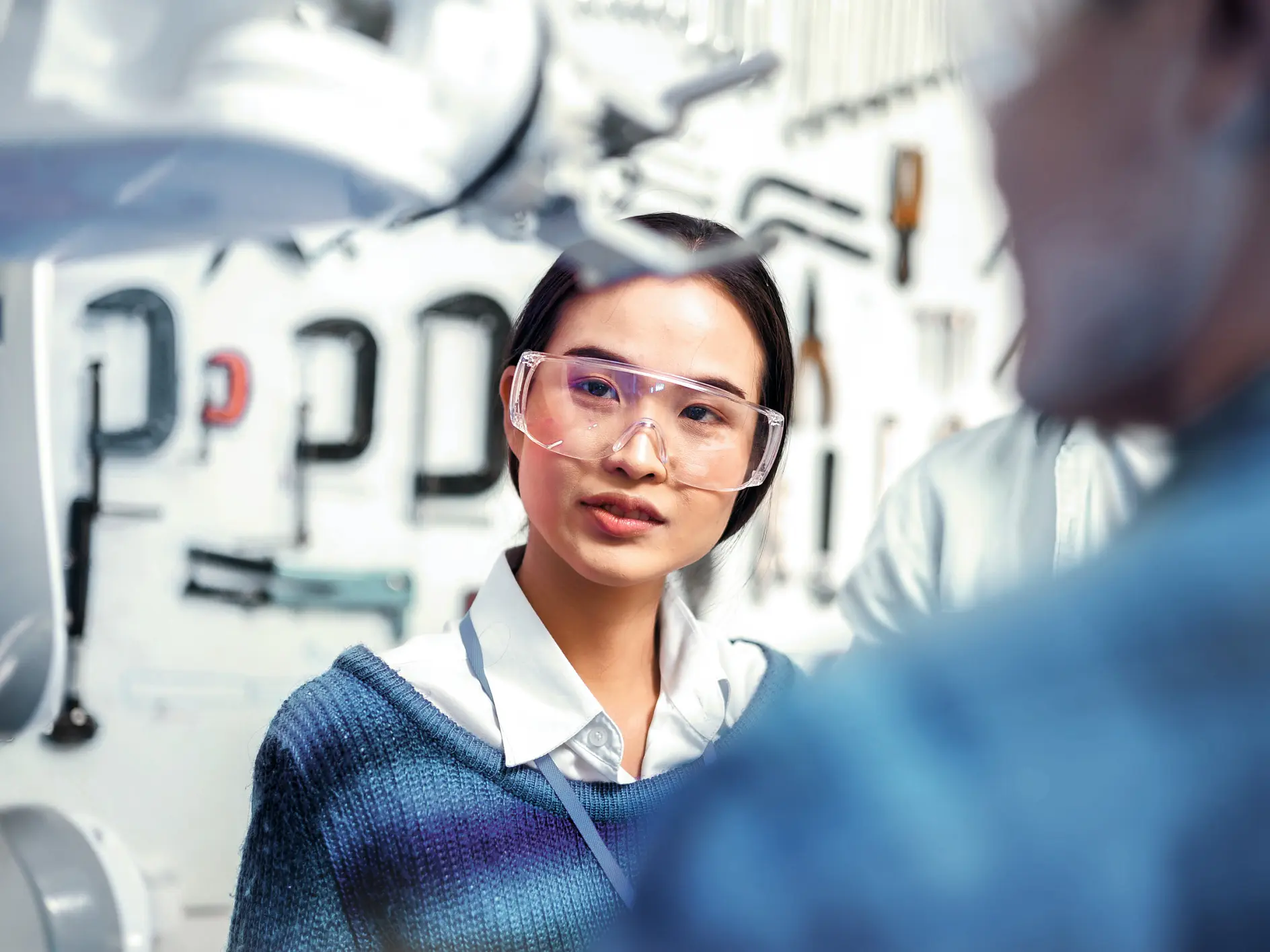 A woman seen from the front and a man visible from behind, both wearing safety glasses, look at a robot. (This text has been generated by AI)
