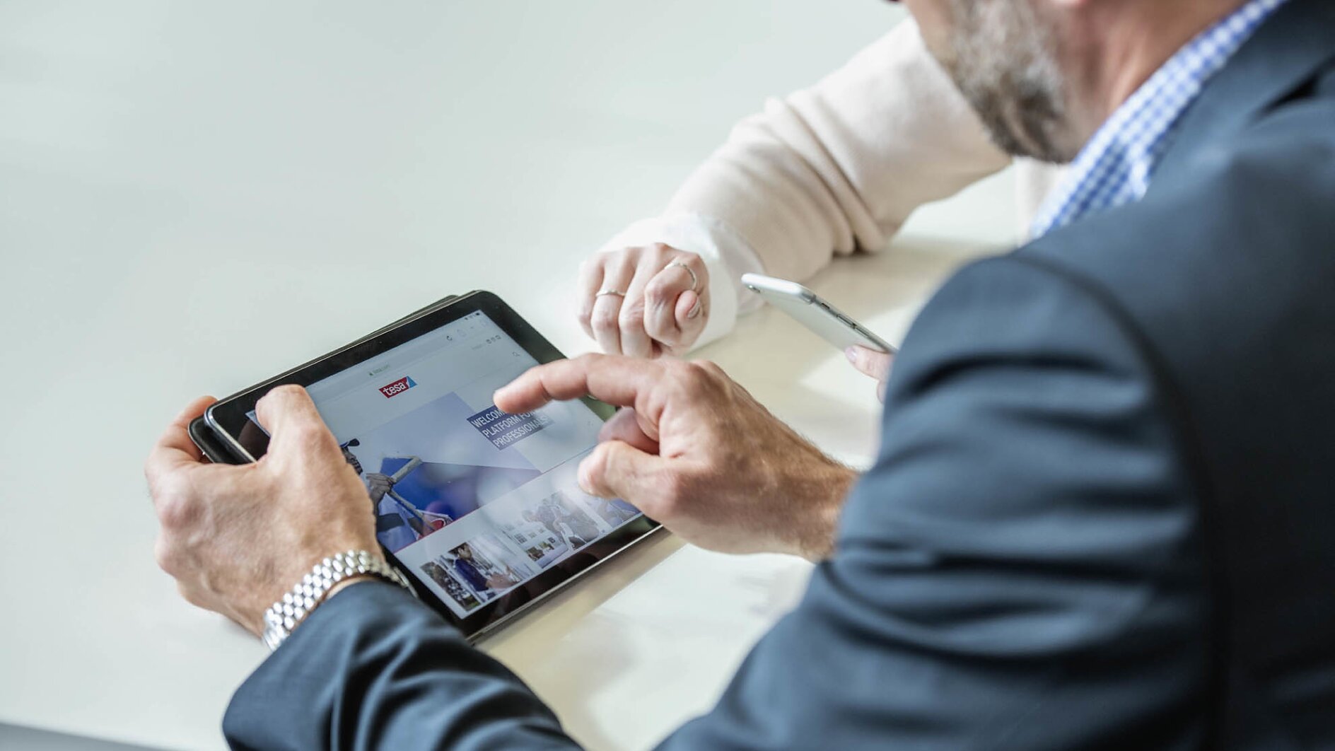 Two people are seated at a table with digital devices. One person in a suit gestures at a tablet screen displaying a website, while the other person holds a smartphone. Their faces are not visible. (This text has been generated by AI)