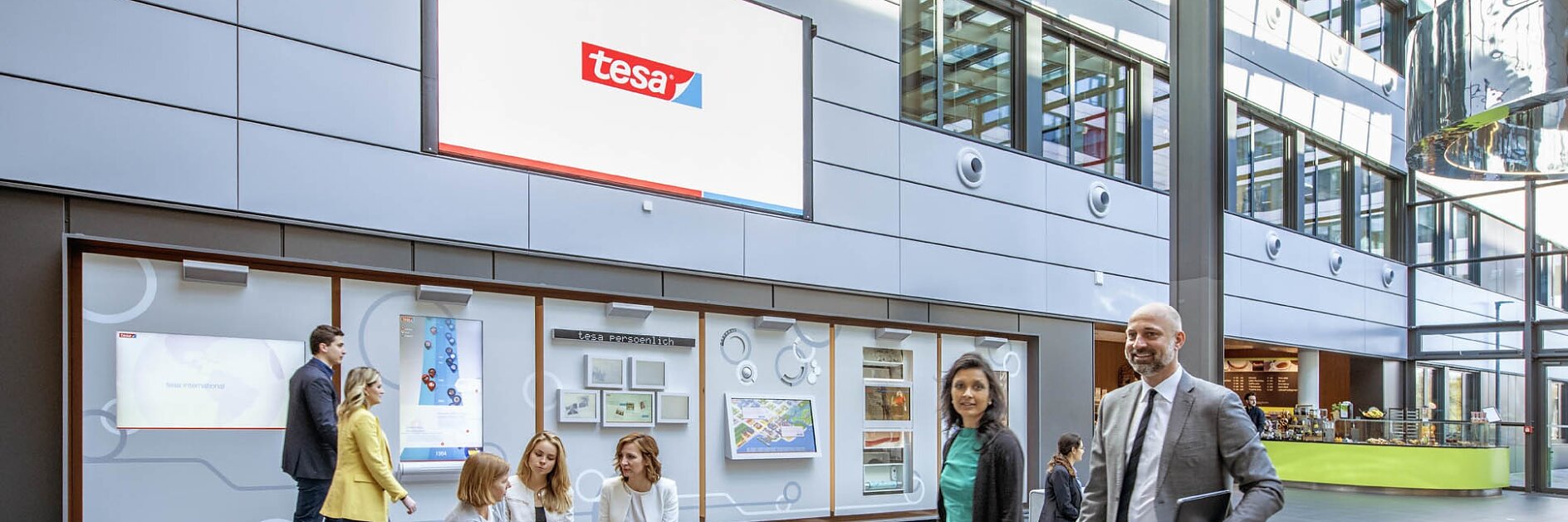 Several people in business attire are in a modern office lobby. A large digital screen displays the logo tesa. Some individuals are seated on modular furniture, while others walk across the polished floor. The space is bright with natural light. (This text has been generated by AI)