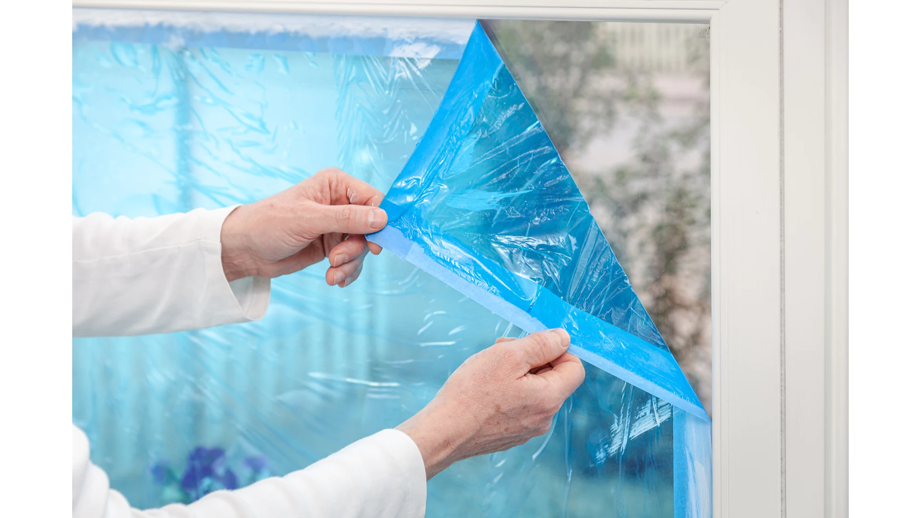 Eine Person zieht blaue Tesafilm-Schutzfolie von einer Fensterscheibe ab. Die Person trägt ein weißes langärmeliges Kleidungsstück, und der Fensterrahmen ist weiß. Außerhalb des Fensters sind unscharfe Pflanzen zu sehen. (This text has been generated by AI)