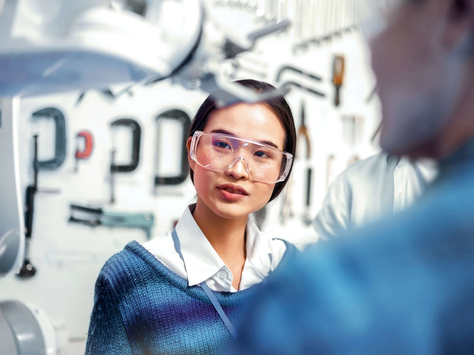 Eine Person, die eine Schutzbrille und einen blauen Pullover trägt, konzentriert sich auf eine Aufgabe. Im Hintergrund sind Werkzeuge und Geräte an einer Wand zu sehen. Es scheint sich um ein Labor oder eine Werkstatt zu handeln, in der Tesafilmrollen eine wichtige Rolle spielen. (This text has been generated by AI)