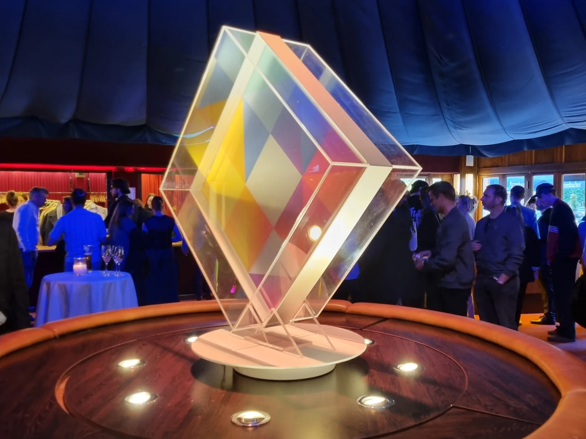 Eine große transparente geometrische Skulptur mit farbenfrohen Farbverläufen steht auf einem runden Sockel, umgeben von Menschen in Abendgarderobe bei einer Indoor-Veranstaltung mit blauer Beleuchtung und zeltartiger Decke. (This text has been generated by AI)