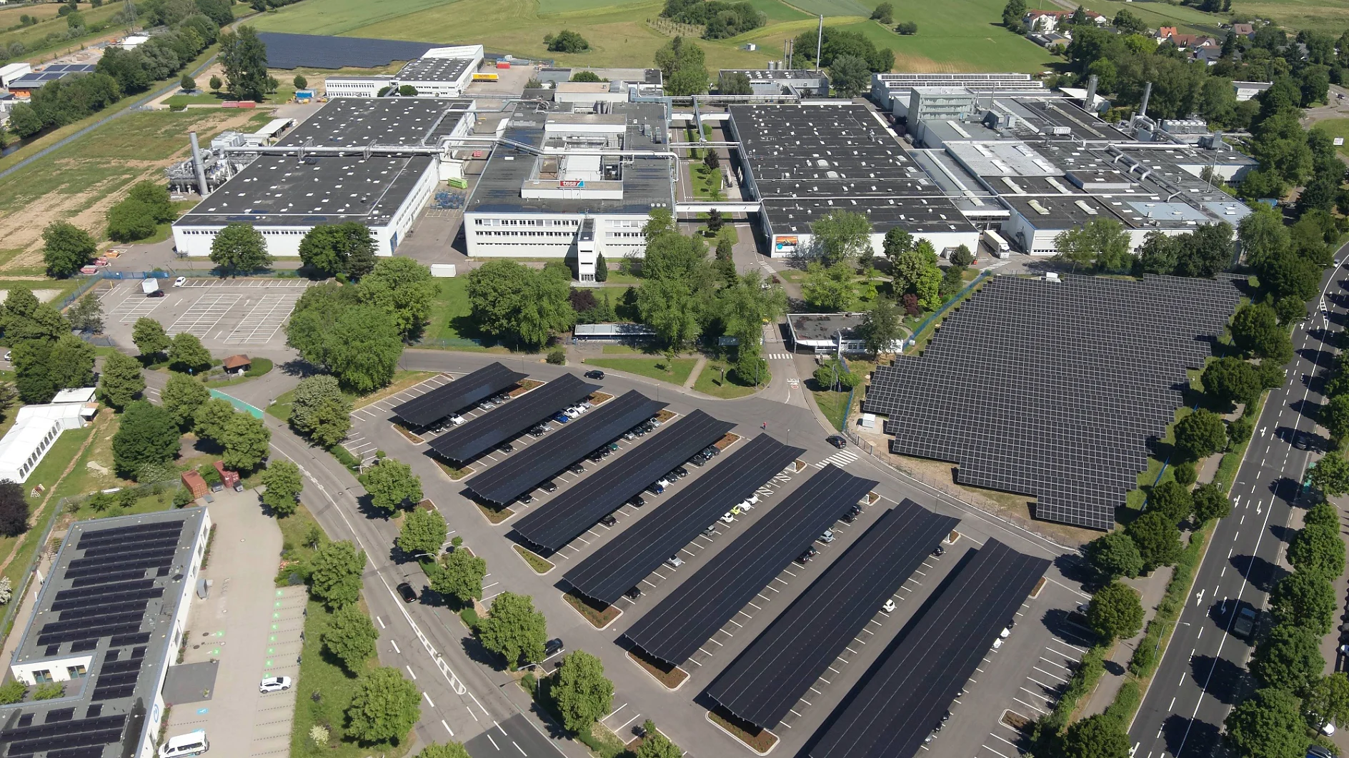 Luftaufnahme eines großen Industriekomplexes mit mehreren Gebäuden, deren Dächer mit Tesa-Klebeband befestigte Solarmodule aufweisen, einem Parkplatz mit Solarmoduldächern, umgebender Grünanlage und einem Wohngebiet in der Ferne. (This text has been generated by AI)