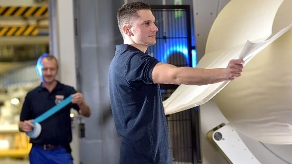 Zwei Männer in Arbeitsuniformen bedienen Maschinen in einer Industrieanlage. Ein Mann prüft eine große Materialbahn, die aus einer Maschine kommt, während der andere im Hintergrund eine Rolle blaues Tesa-Klebeband in der Hand hält. (This text has been generated by AI)