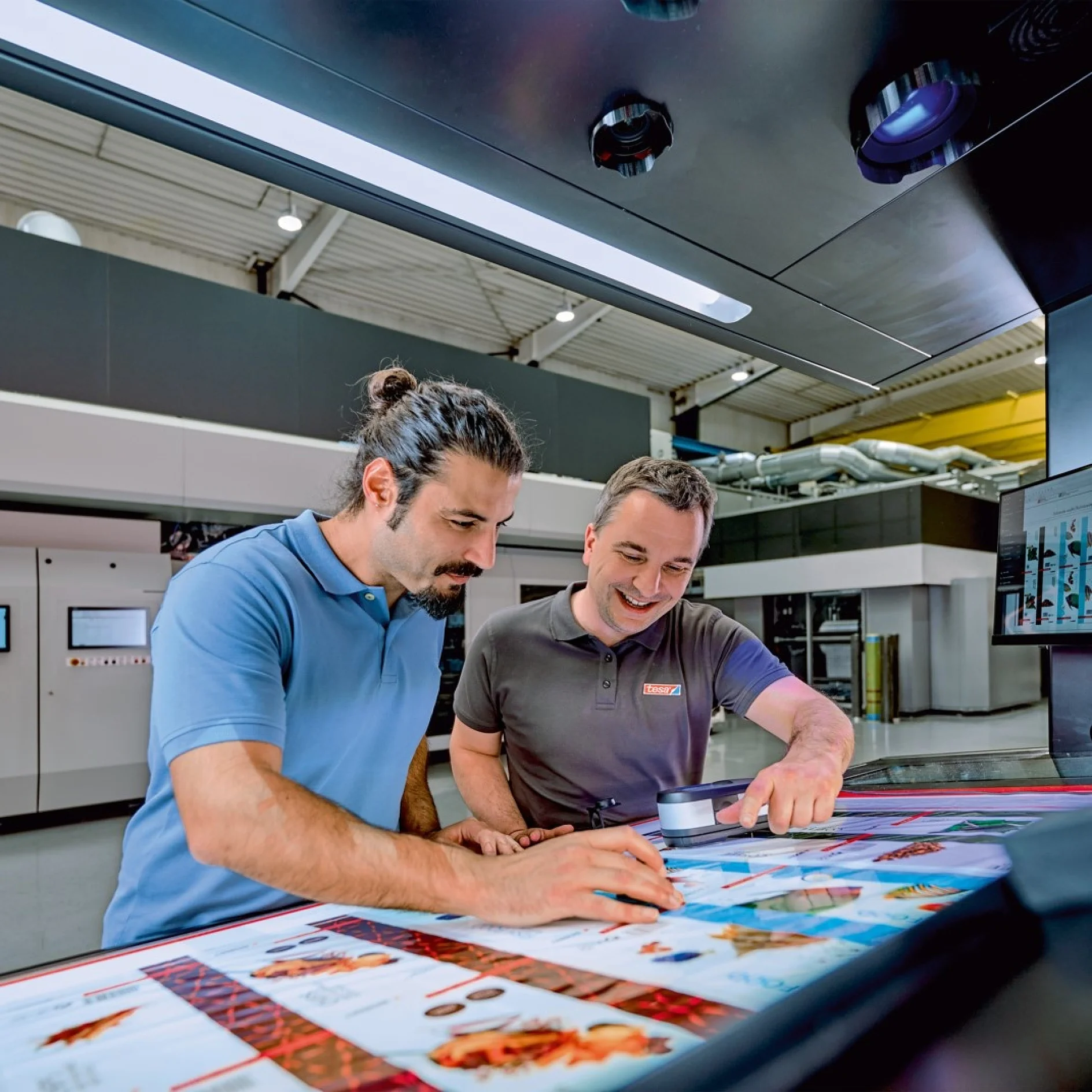 Two men stand at a large digital screen in an industrial setting, examining colorful printed materials and discussing the images while touching the screen. Printing equipment and machinery are visible in the background, with tesa tape featured prominently in the workspace. (This text has been generated by AI)