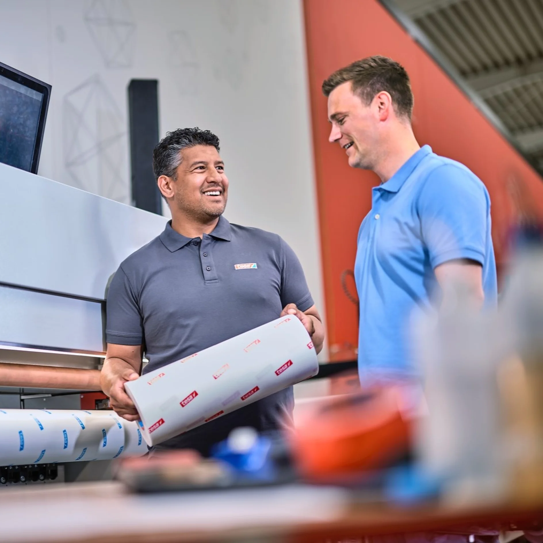 Two men standing indoors, one holding a roll of printed material with tesa tape. They are smiling and talking in a workspace with a large machine and equipment visible in the background. (This text has been generated by AI)