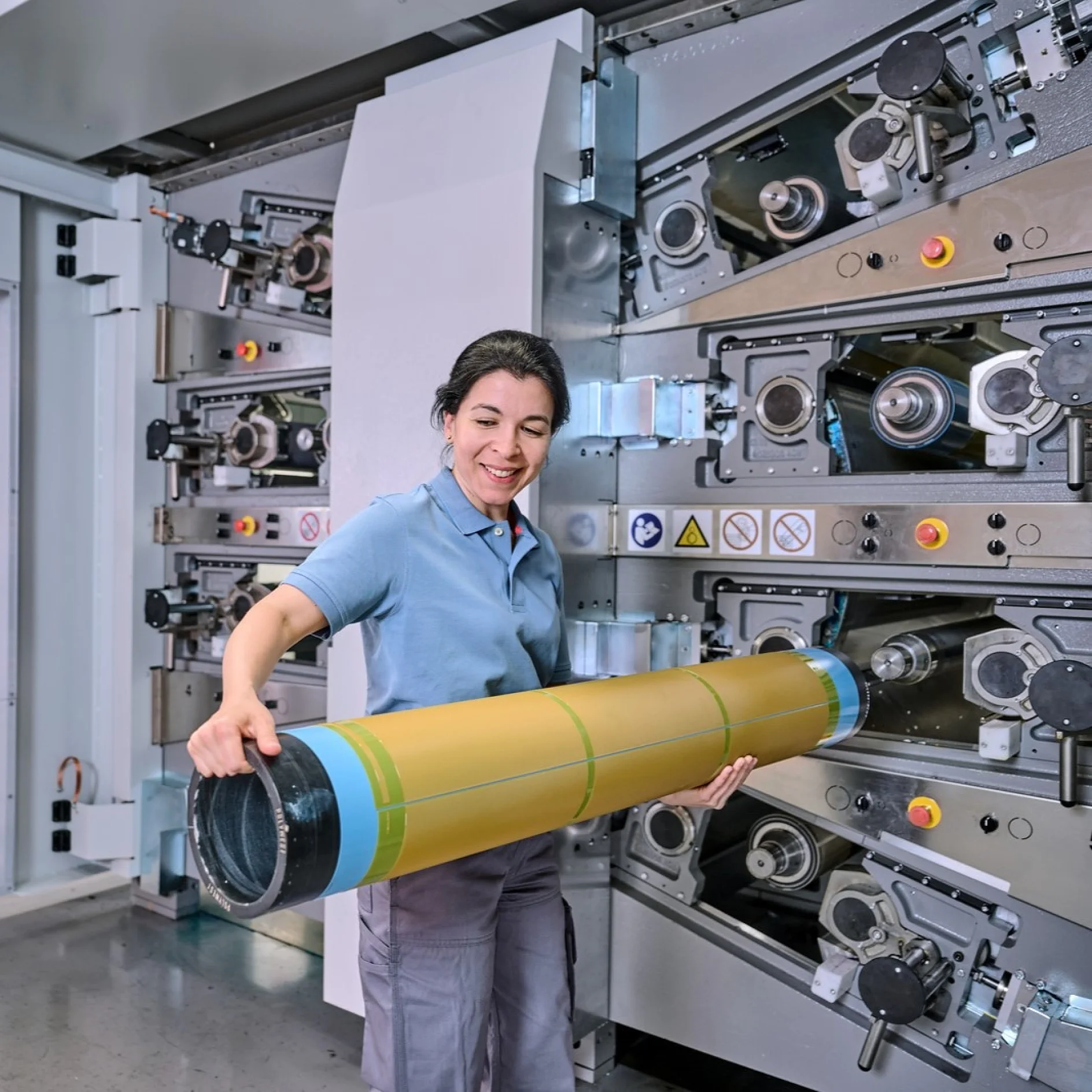 A woman in a polo shirt stands in front of industrial printing machinery, smiling while holding a large yellow and green cylindrical tesa tape roller. The machine behind her has various gears, tesa tape rollers, and control buttons. (This text has been generated by AI)