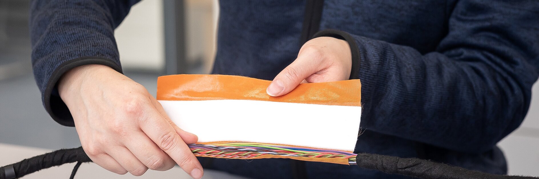 Eine Person in einer tesa Electrical Systems Jacke hält ein Stück orangefarbenes tesa Klebeband über farbcodierte elektrische Drähte auf einem Tisch. (This text has been generated by AI)
