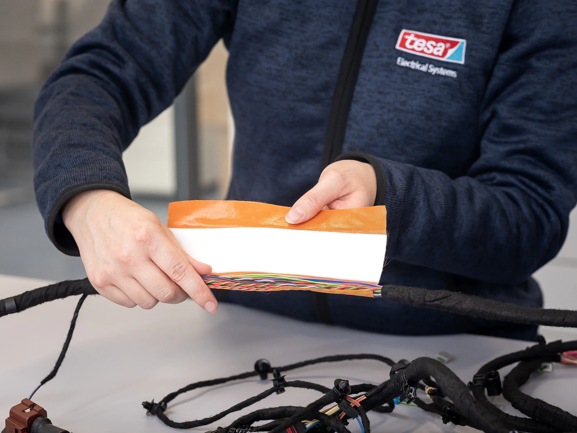 Eine Person in einer tesa Electrical Systems Jacke hält ein Stück orangefarbenes tesa Klebeband über farbcodierte elektrische Drähte auf einem Tisch. (This text has been generated by AI)