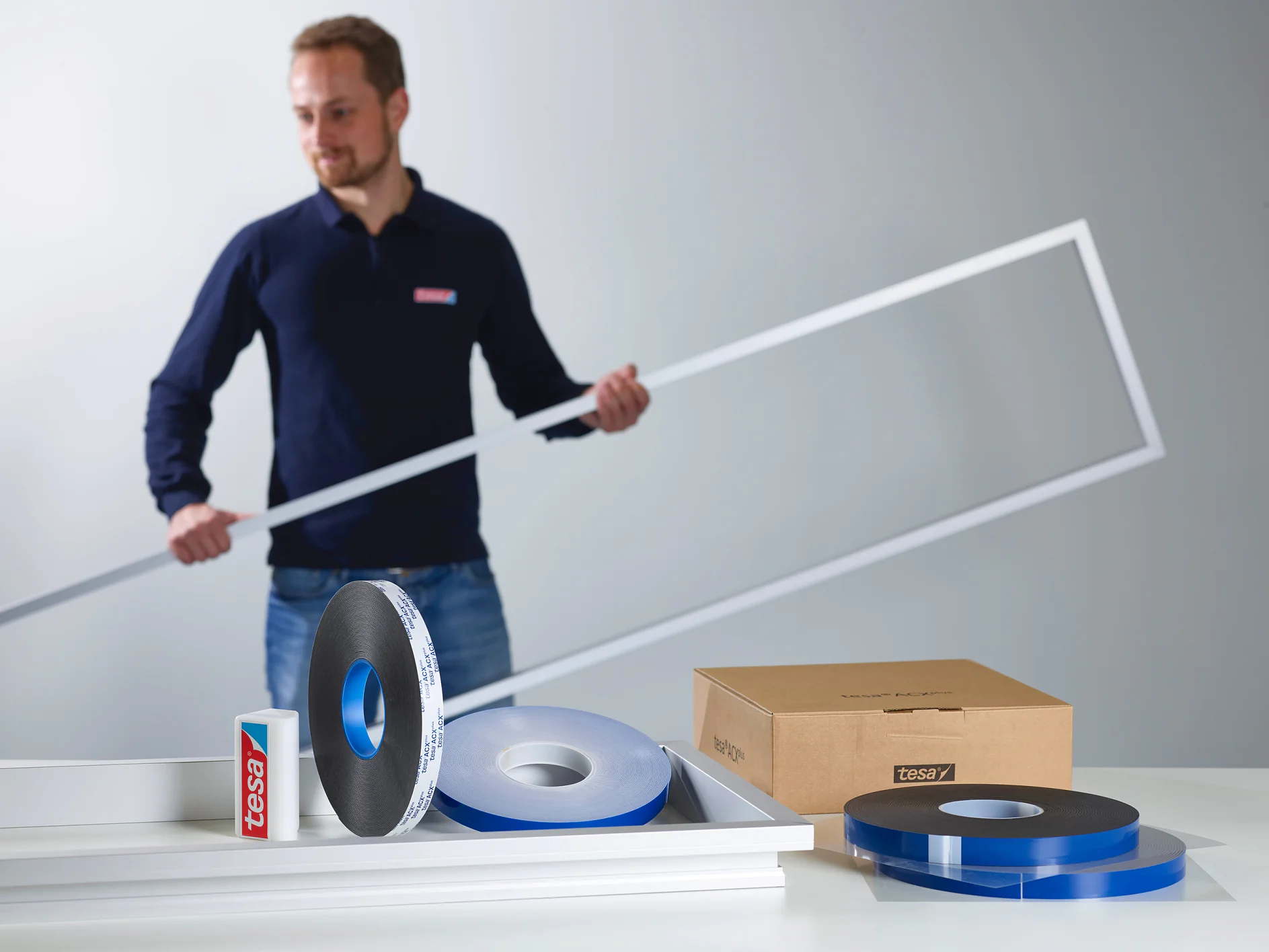 Ein Mann in einem marineblauen Hemd hält einen großen Rahmen. Auf einem weißen Tisch liegen Rollen mit Tesaband, eine Tesabandbox, ein Karton und Aluminiumrahmenstücke. (This text has been generated by AI)