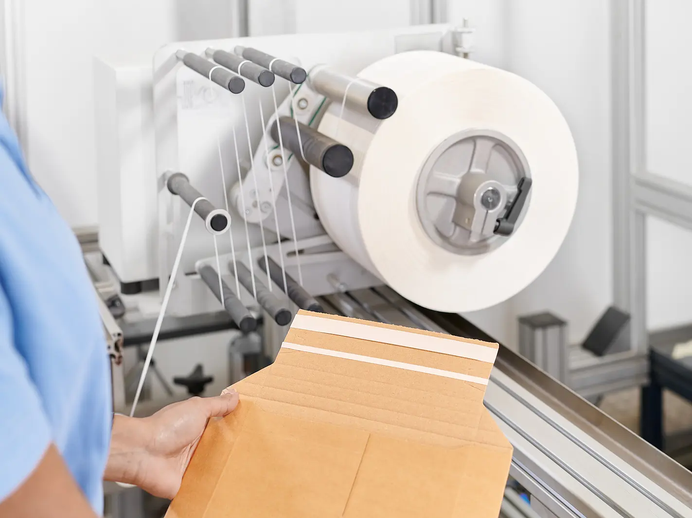 A person holds a flat brown envelope with adhesive strips in front of a machine dispensing a large roll of white tesa tape in an industrial setting.