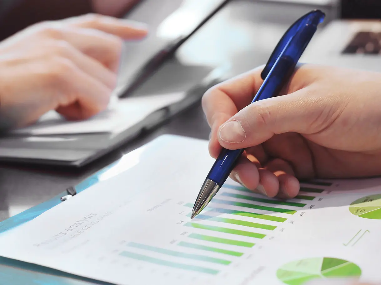 A person holds a blue pen over a printed sheet with green bar and pie charts secured with tesa tape, while another person points at a laptop in the background. The focus is on business data analysis.