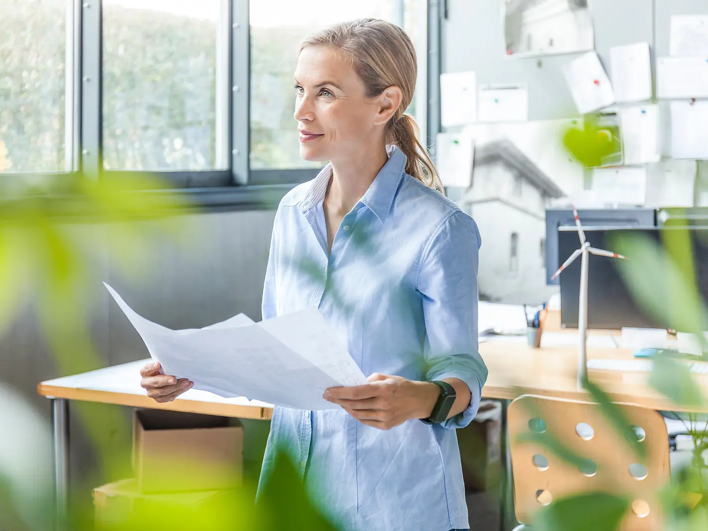 A woman in a light blue shirt stands in an office holding papers. She faces left, and the workspace behind her includes a desk, chair, computer, and papers attached to the wall with tesa tape. Green plants are visible in the foreground.