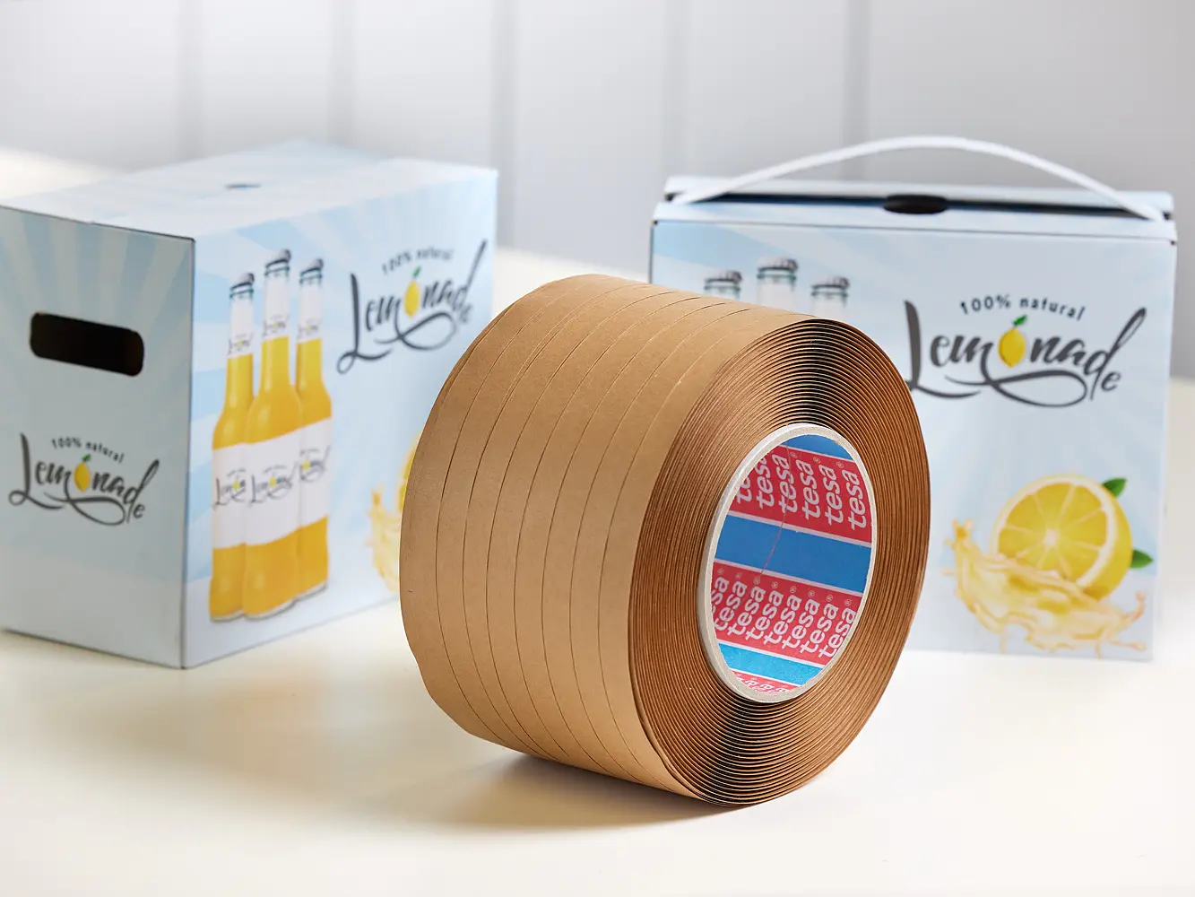 A large roll of brown tesa tape stands in front of two cardboard boxes labeled lemonade with images of lemonade bottles and lemons on a white background.