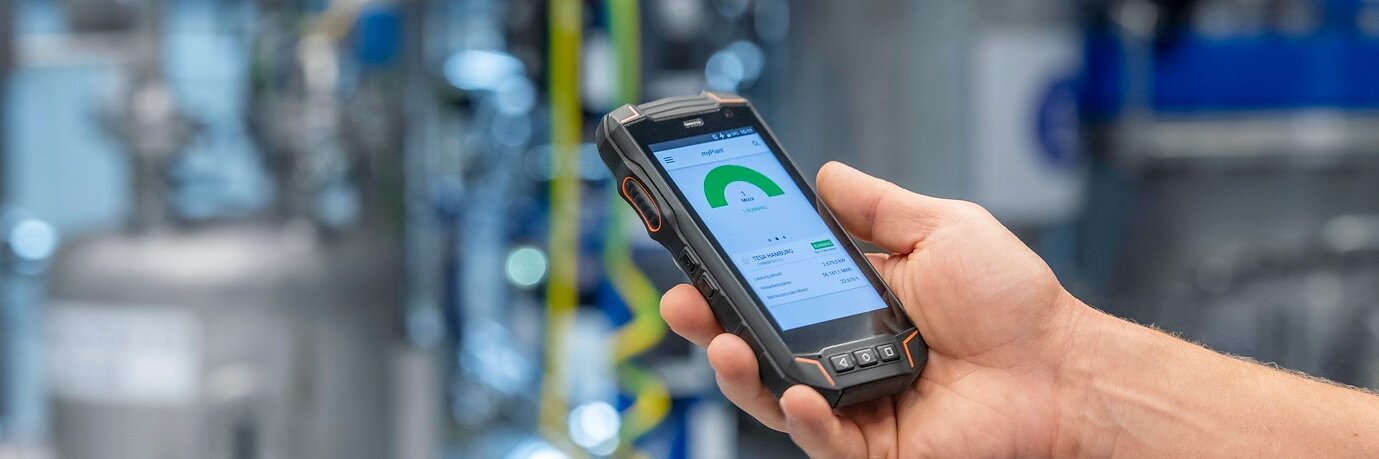 Eine Person hält ein robustes Handgerät mit einer grünen Anzeige und eingeblendeten Daten in einer Industrieanlage, in der tesa-Klebeband an Rohren angebracht ist. (This text has been generated by AI)