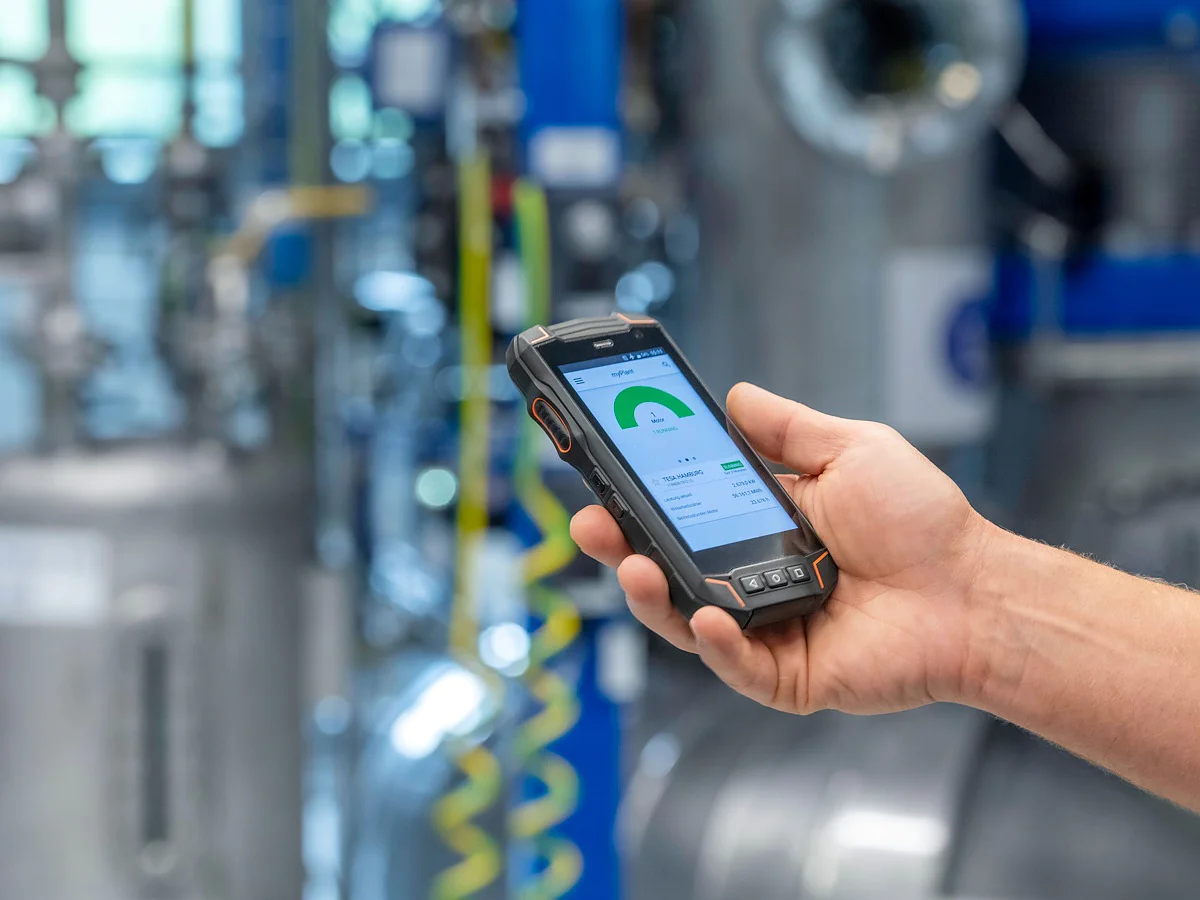 Eine Person hält ein robustes Handgerät mit einer grünen Anzeige und eingeblendeten Daten in einer Industrieanlage, in der tesa-Klebeband an Rohren angebracht ist. (This text has been generated by AI)