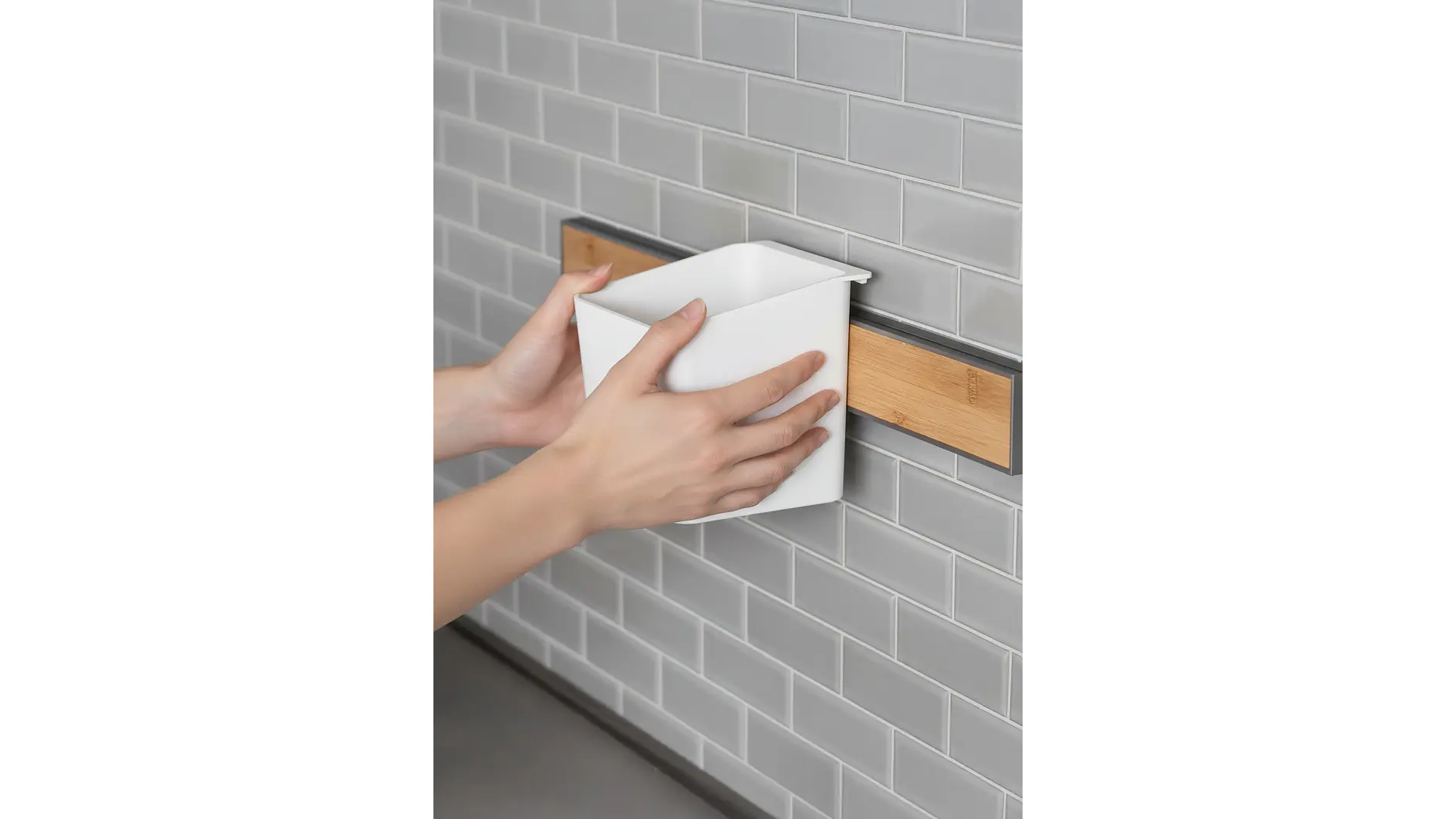 Eine Person befestigt einen weißen rechteckigen Behälter auf einer Holzleiste, die mit Tesafilm an einer grauen Kachelwand befestigt ist. (This text has been generated by AI)