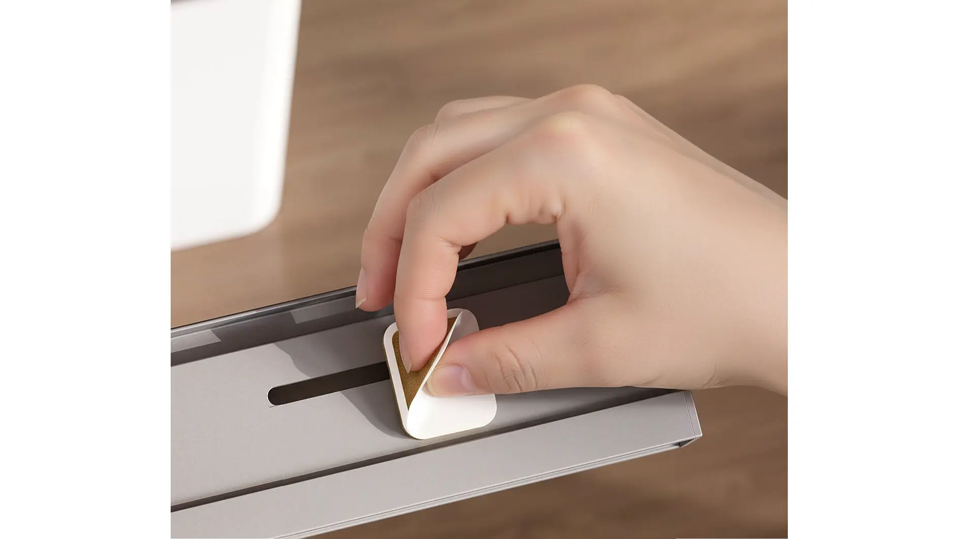 Eine Hand bringt einen kleinen, weißen, quadratischen Klebehaken mit Tesafilm auf einer grauen Metallfläche an. Der Hintergrund ist unscharf. (This text has been generated by AI)