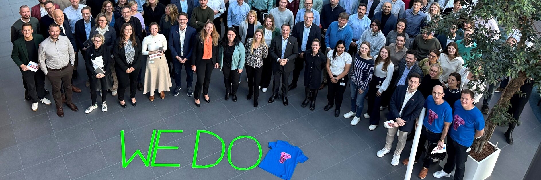A large group poses indoors for a photo; a blue T-shirt and "WEDO" made with green tesa tape are on the floor. Some hold papers. (This text has been generated by AI)