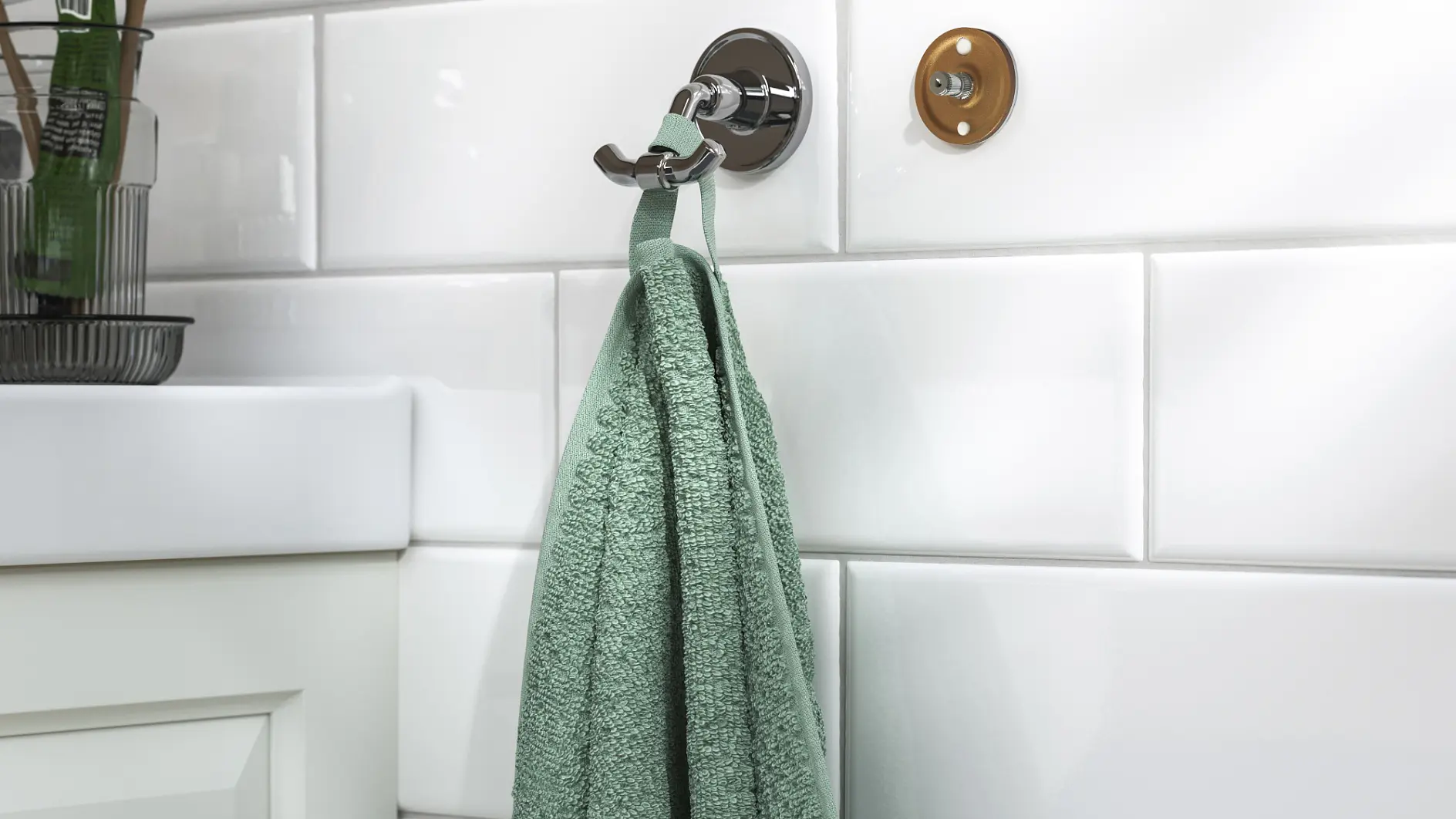 Ein grünes Handtuch hängt an einem mit Tesafilm befestigten Metallhaken an einer weiß gefliesten Badezimmerwand. In der Nähe befinden sich ein Waschbecken und ein Schrank.