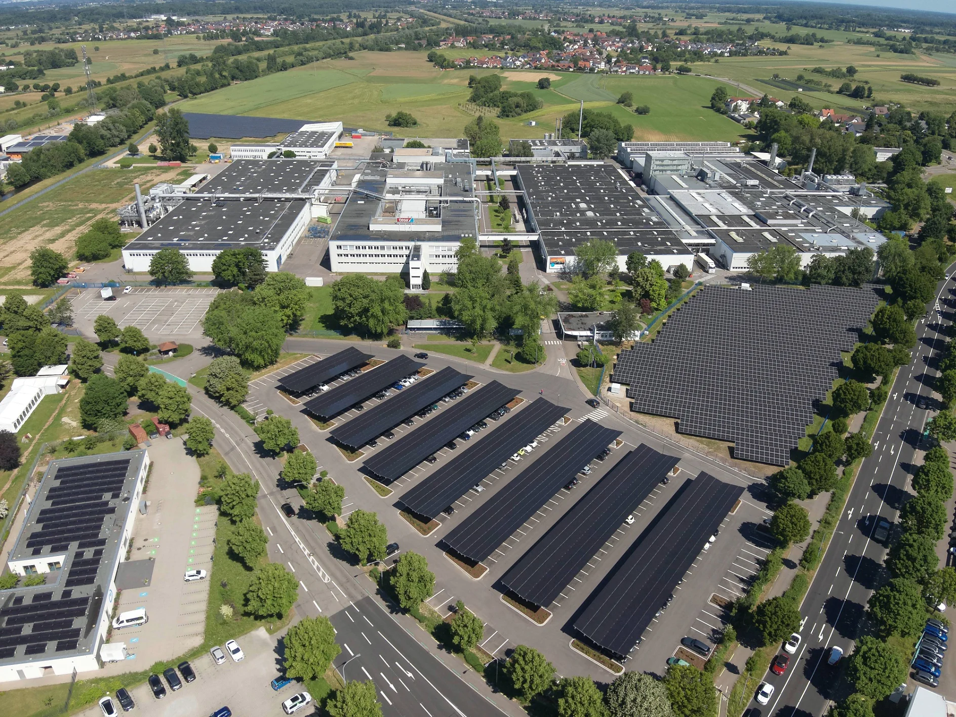 Luftaufnahme eines Industriekomplexes mit Solarzellen auf den Dächern und Parkplätzen, umgeben von Feldern und einer nahe gelegenen Stadt. (This text has been generated by AI)