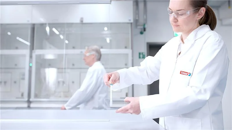 A person in a white lab coat and safety glasses handles a transparent tesa tape object in a bright lab, another in the background. (This text has been generated by AI)