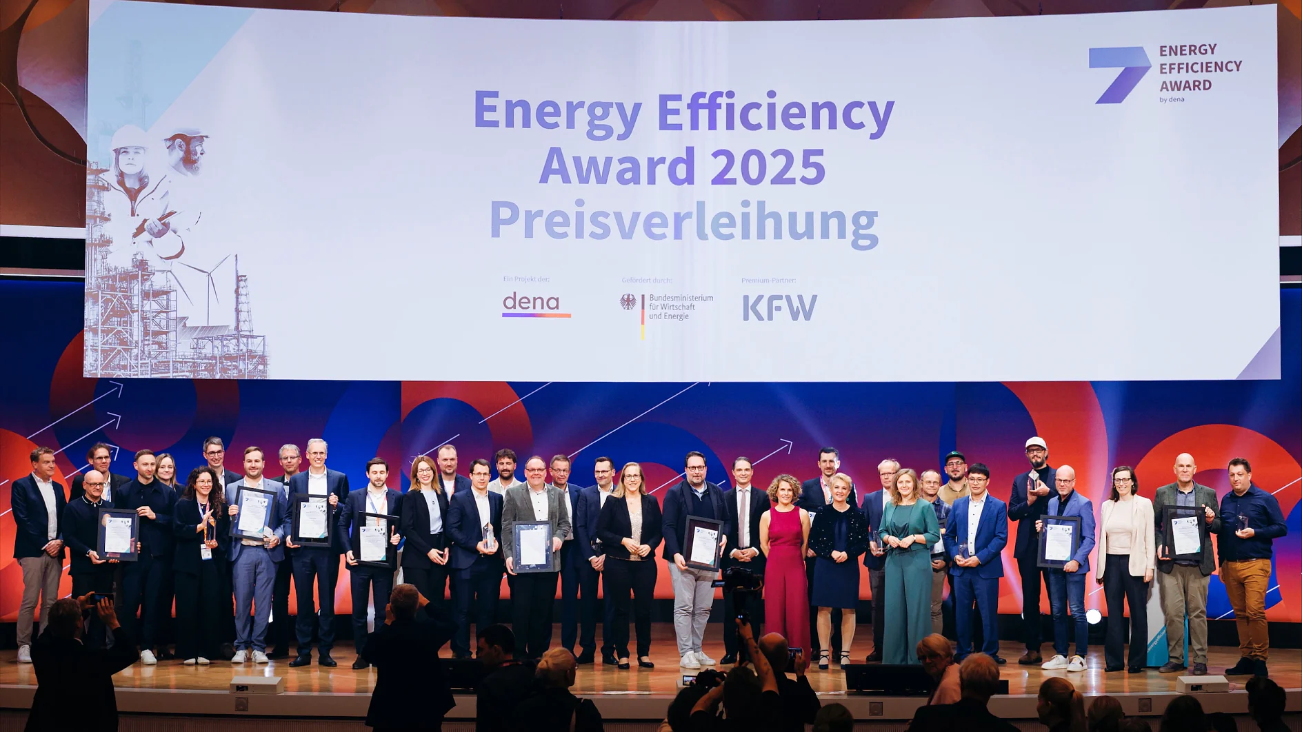 Eine große Gruppe steht auf der Bühne mit Urkunden vor einem Schild des Energy Efficiency Award 2025 und dena-, KFW- und gov-Logos. (This text has been generated by AI)