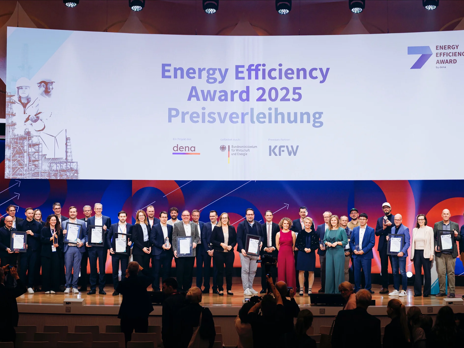 Eine große Gruppe steht auf der Bühne mit Urkunden vor einem Schild des Energy Efficiency Award 2025 und dena-, KFW- und gov-Logos. (This text has been generated by AI)